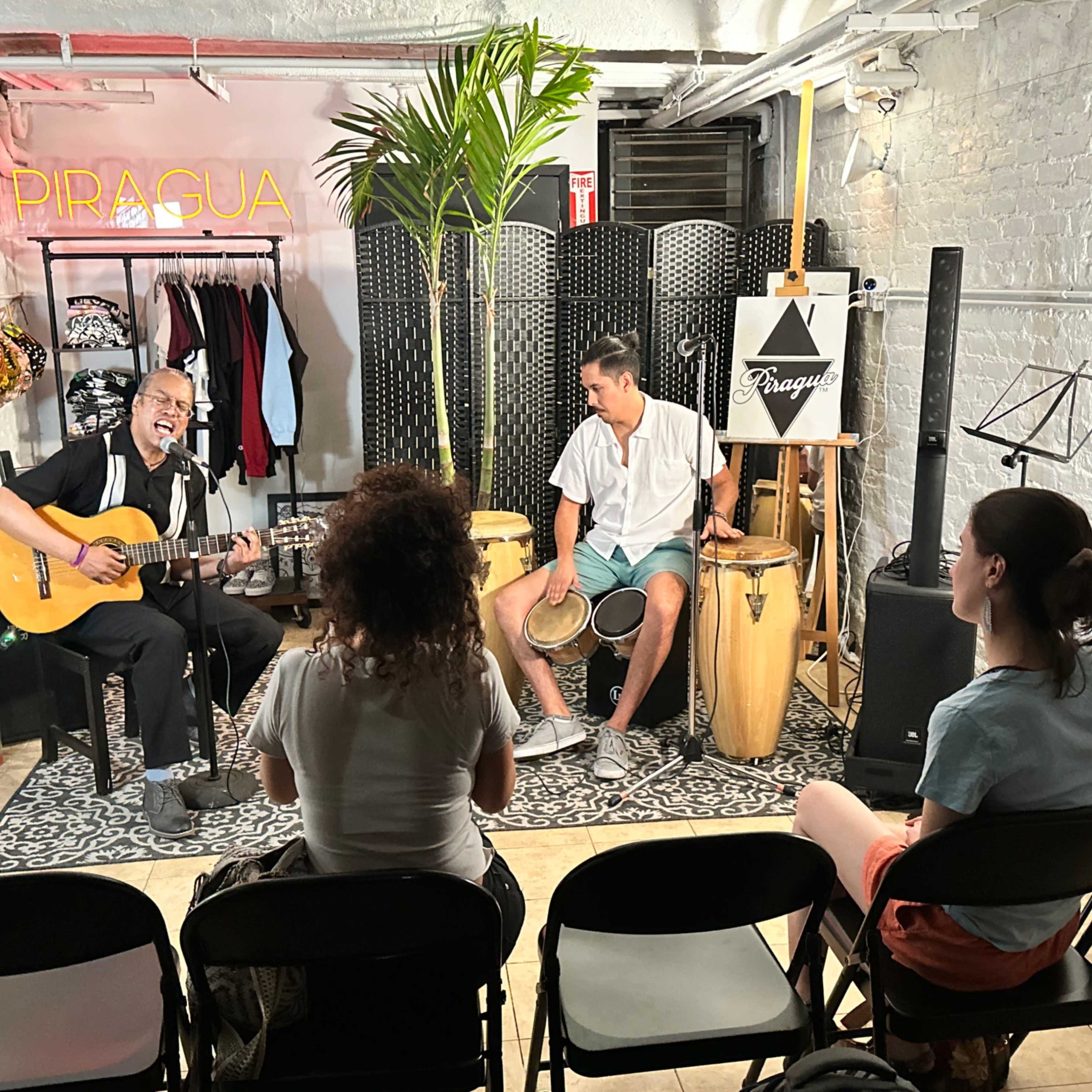 Two musicians perform in a small, lit room with clothing racks and a PIRAGUA sign, while four audience members sit on chairs facing them.