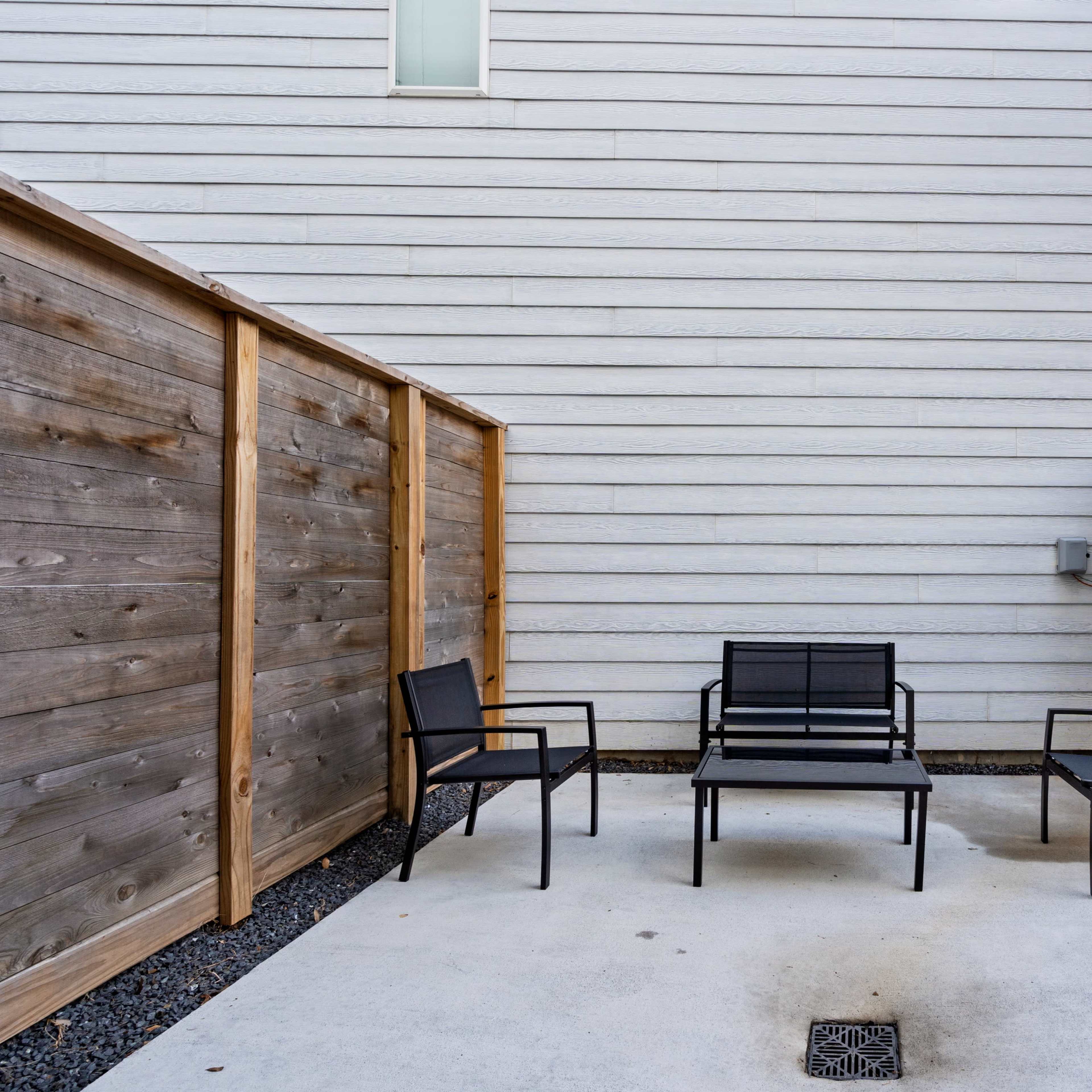 A small patio area features three black metal chairs arranged around a central bench, enclosed by wooden fences on either side.
