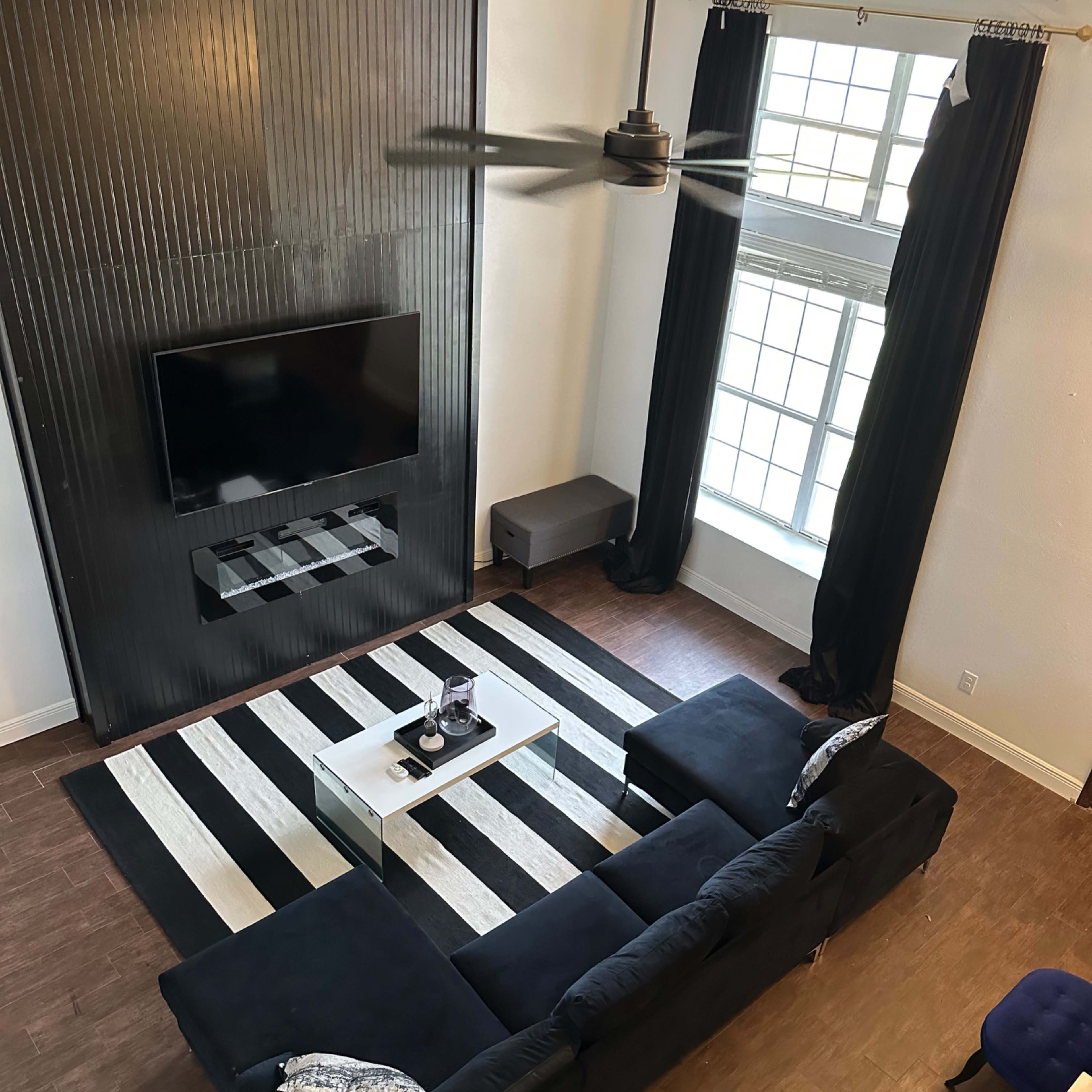A living room with a black and white striped rug, a black sectional sofa, a wall-mounted TV, and large windows dressed with black curtains.