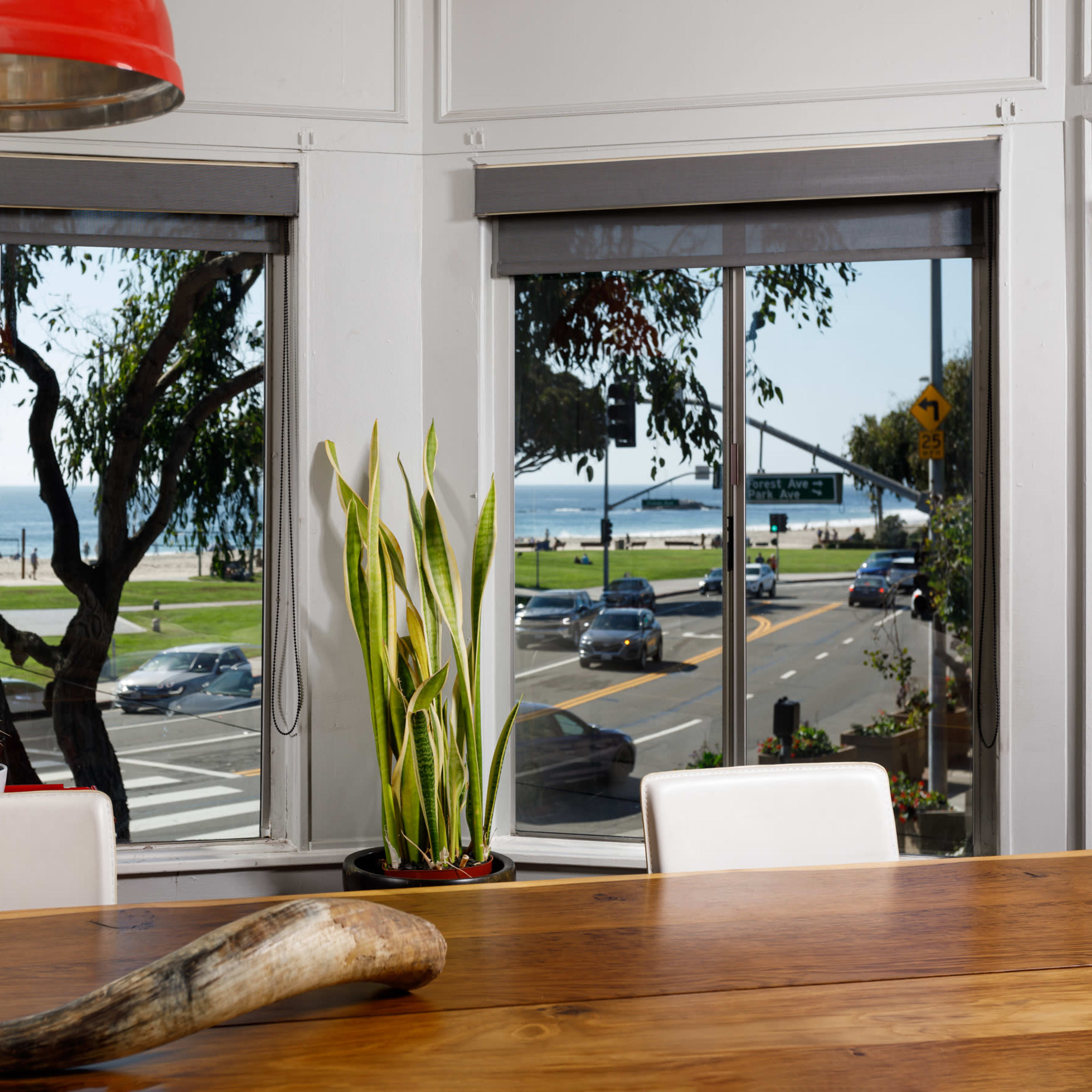 The scene features a dining area with a wooden table, a decorative horn on it, and large windows that provide a view of the ocean and a busy roadway outside.