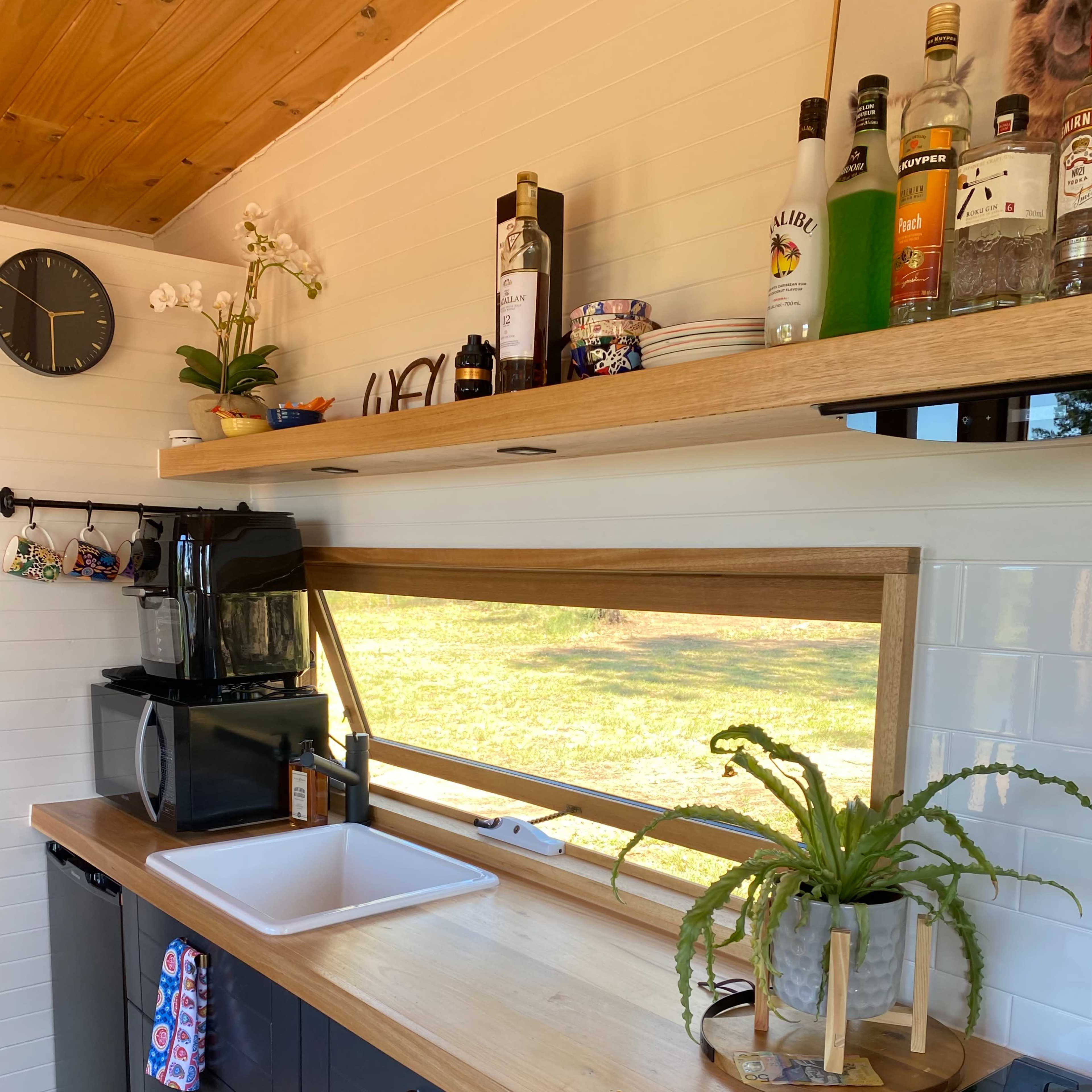 A kitchen with a wooden countertop, a sink, a coffee maker, and a window overlooking an outdoor view, along with shelving displaying drinks and decorative items.