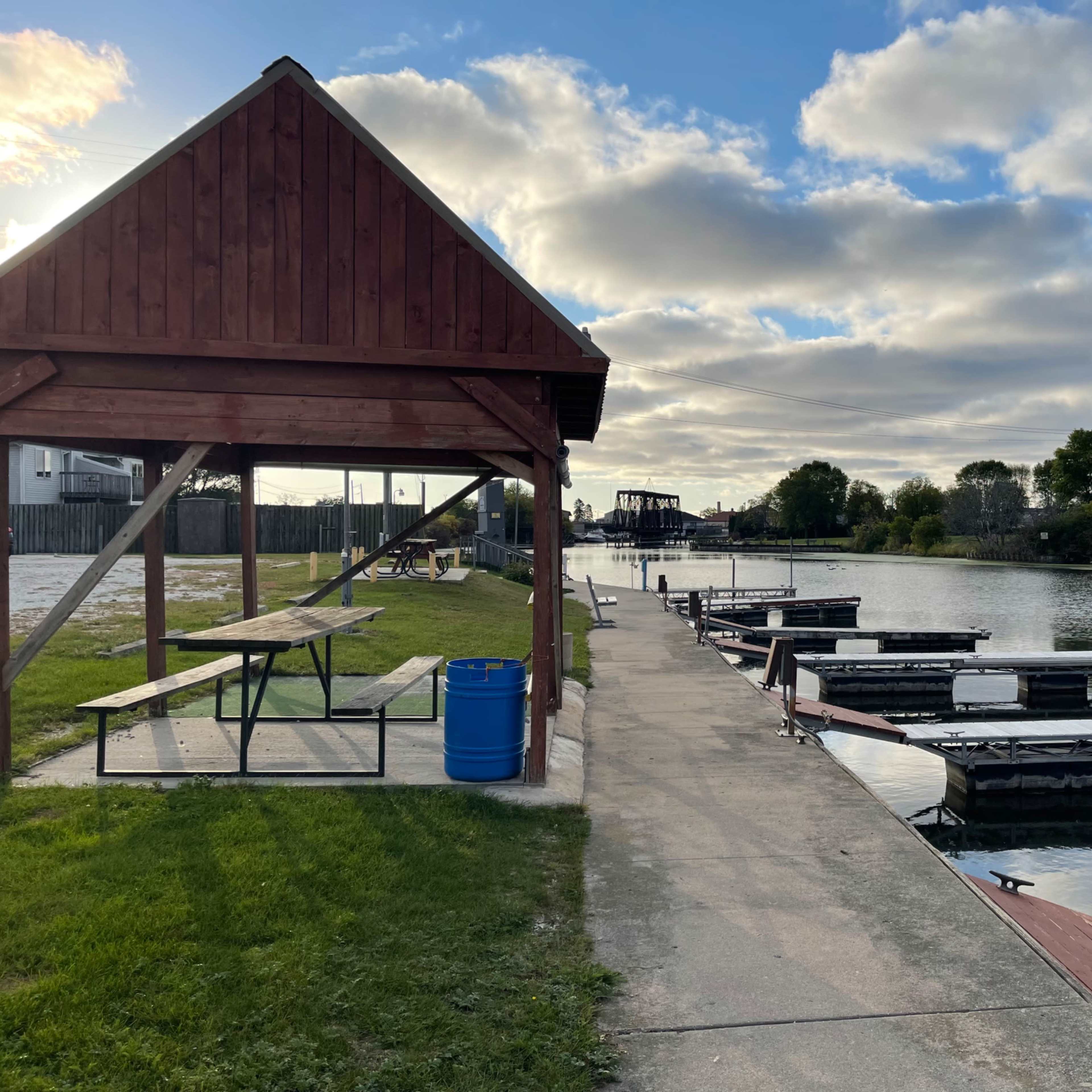 A wooden gazebo alongside a walkway extending to a marina with several docked boats on a calm waterway.