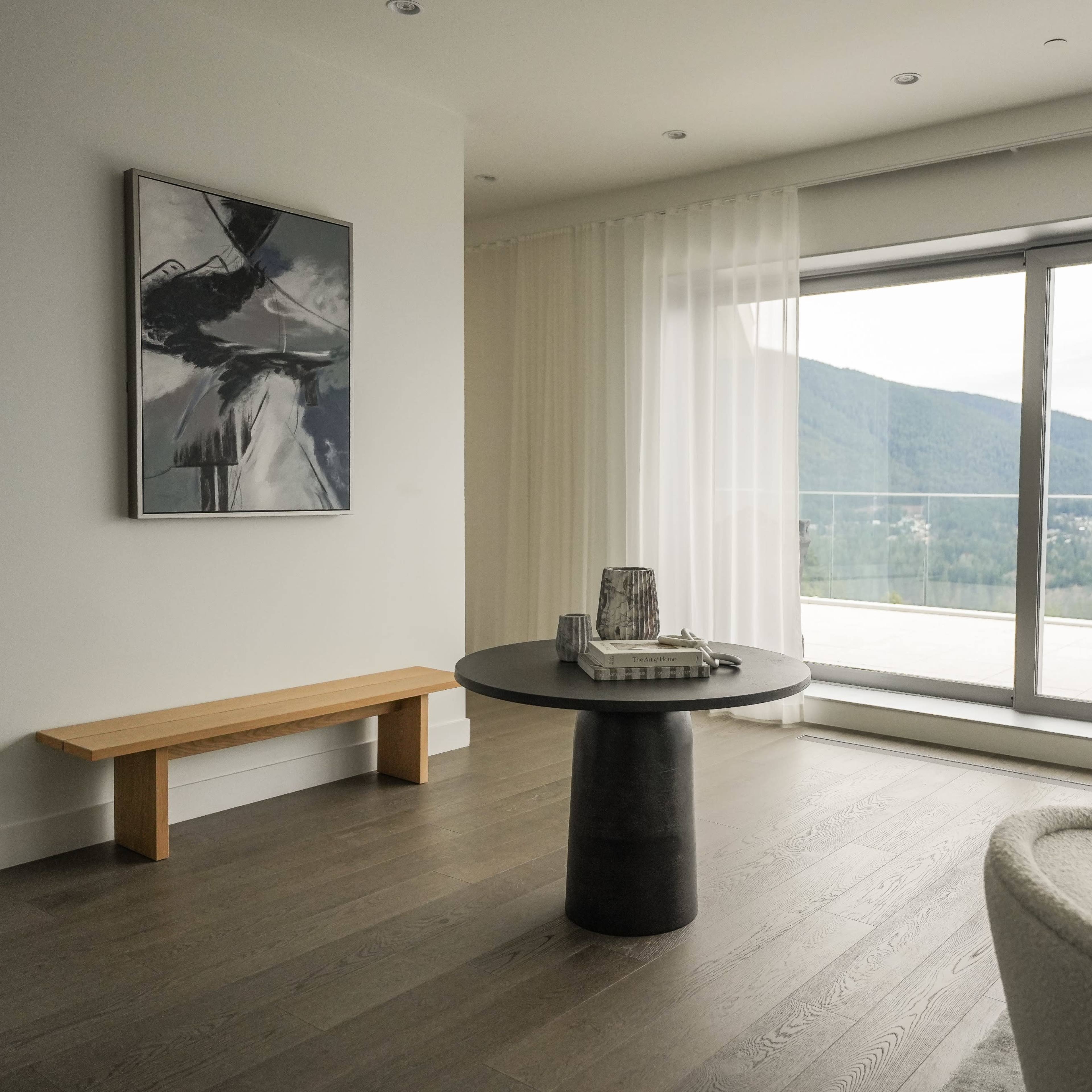 A minimalist living room features a round black table, a wooden bench, and two white upholstered chairs, with large windows offering a view of the mountains.