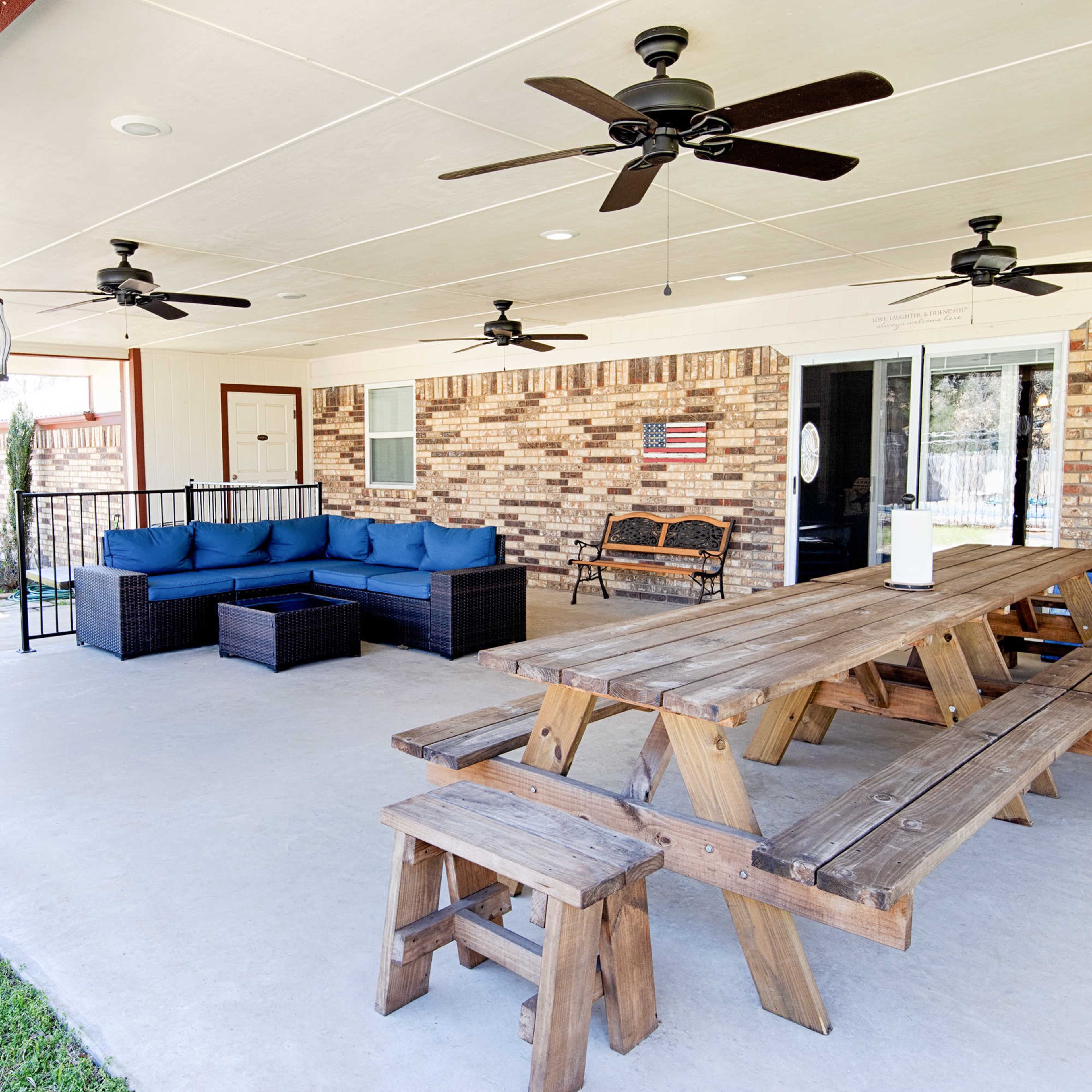 A covered outdoor patio featuring a large wooden dining table, a seating area with blue cushions, and multiple ceiling fans.