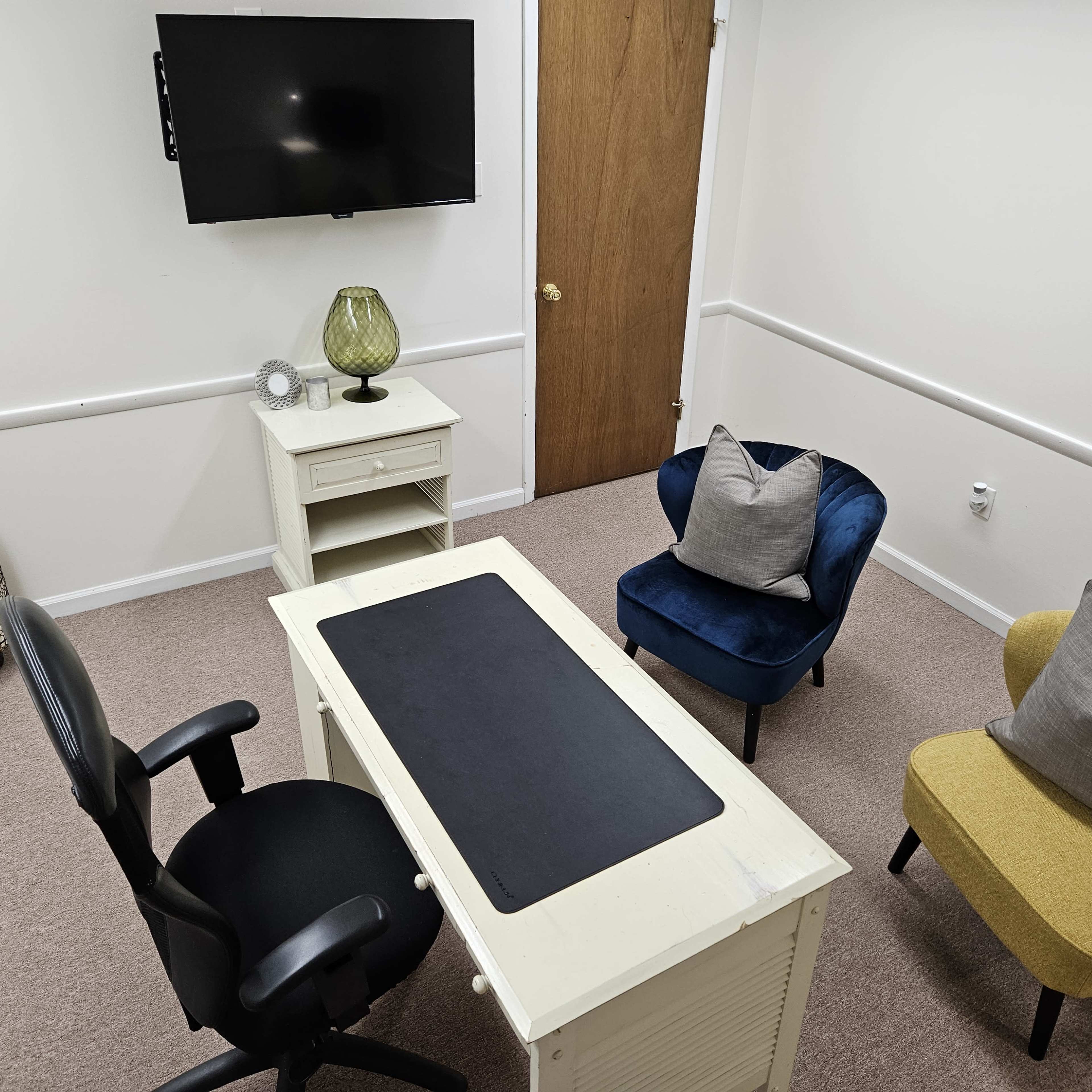 A small office space with a desk, two chairs, a TV mounted on the wall, and a decorative lamp.