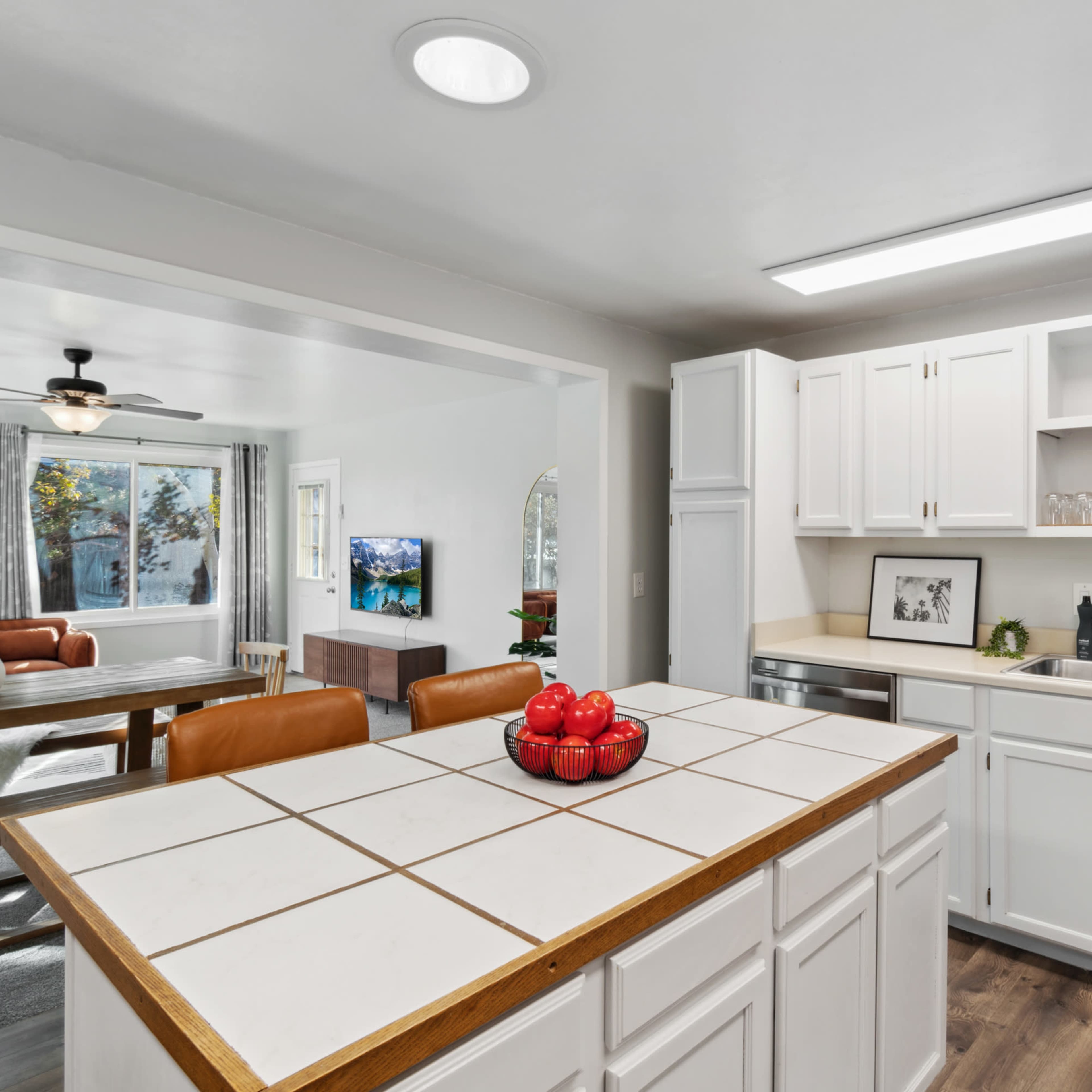 A modern kitchen features white cabinets and countertops with a tile backsplash, overlooking a living room with a television and large windows.