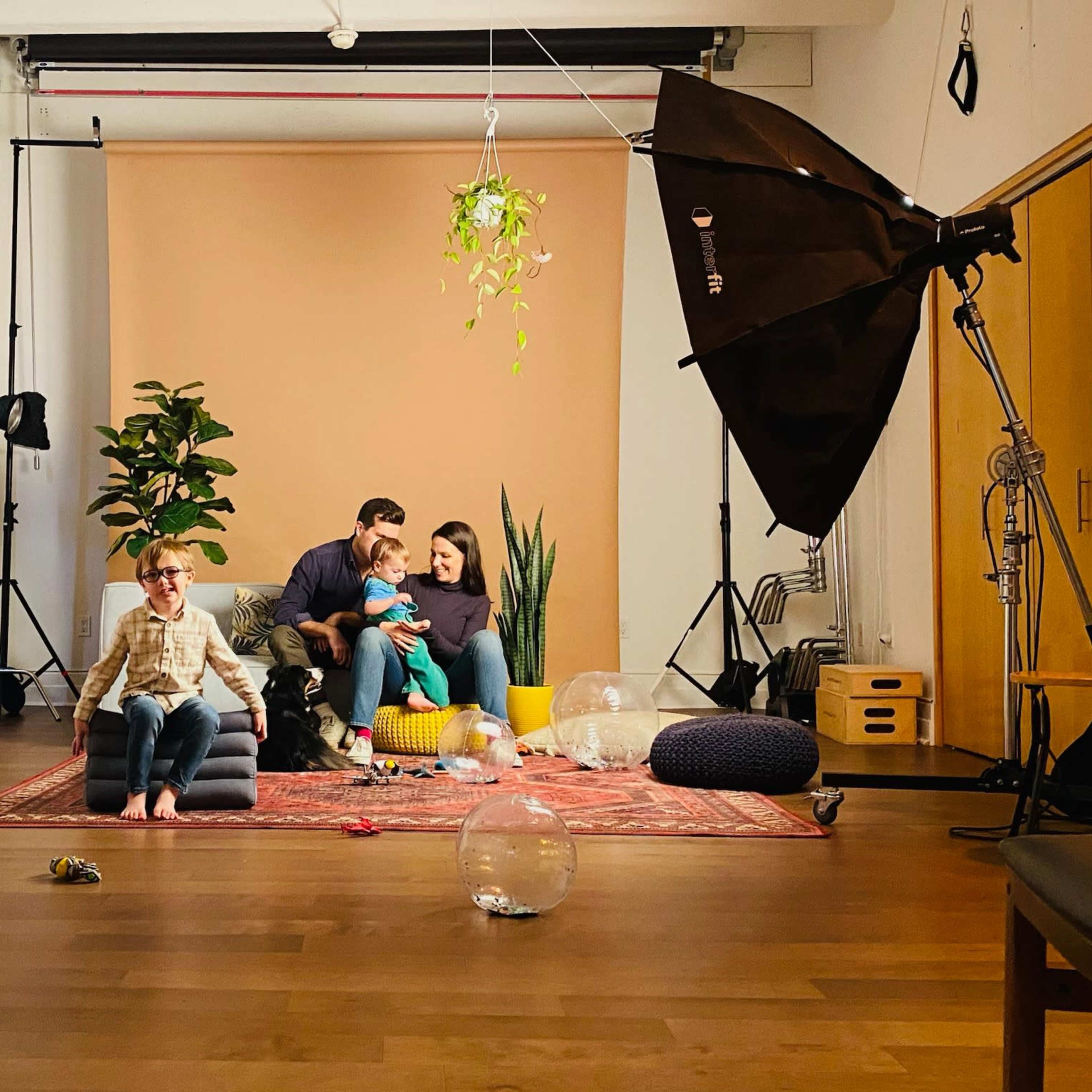 A family of four is seated on a couch in a well-lit photography studio, surrounded by cameras, lighting equipment, and various props.