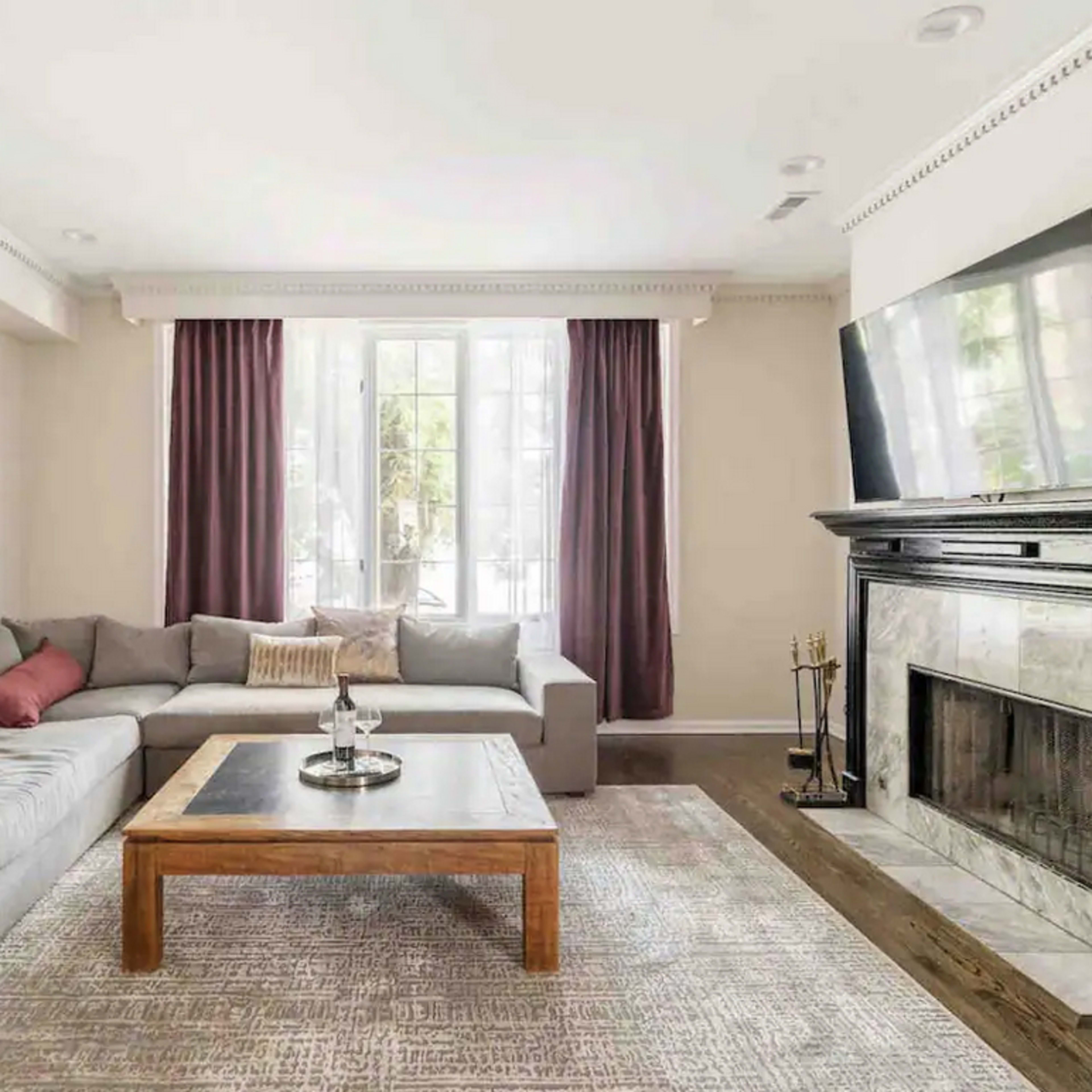 A modern living room features a gray sectional sofa, a wooden coffee table, and a marble fireplace, with large windows dressed in light curtains.