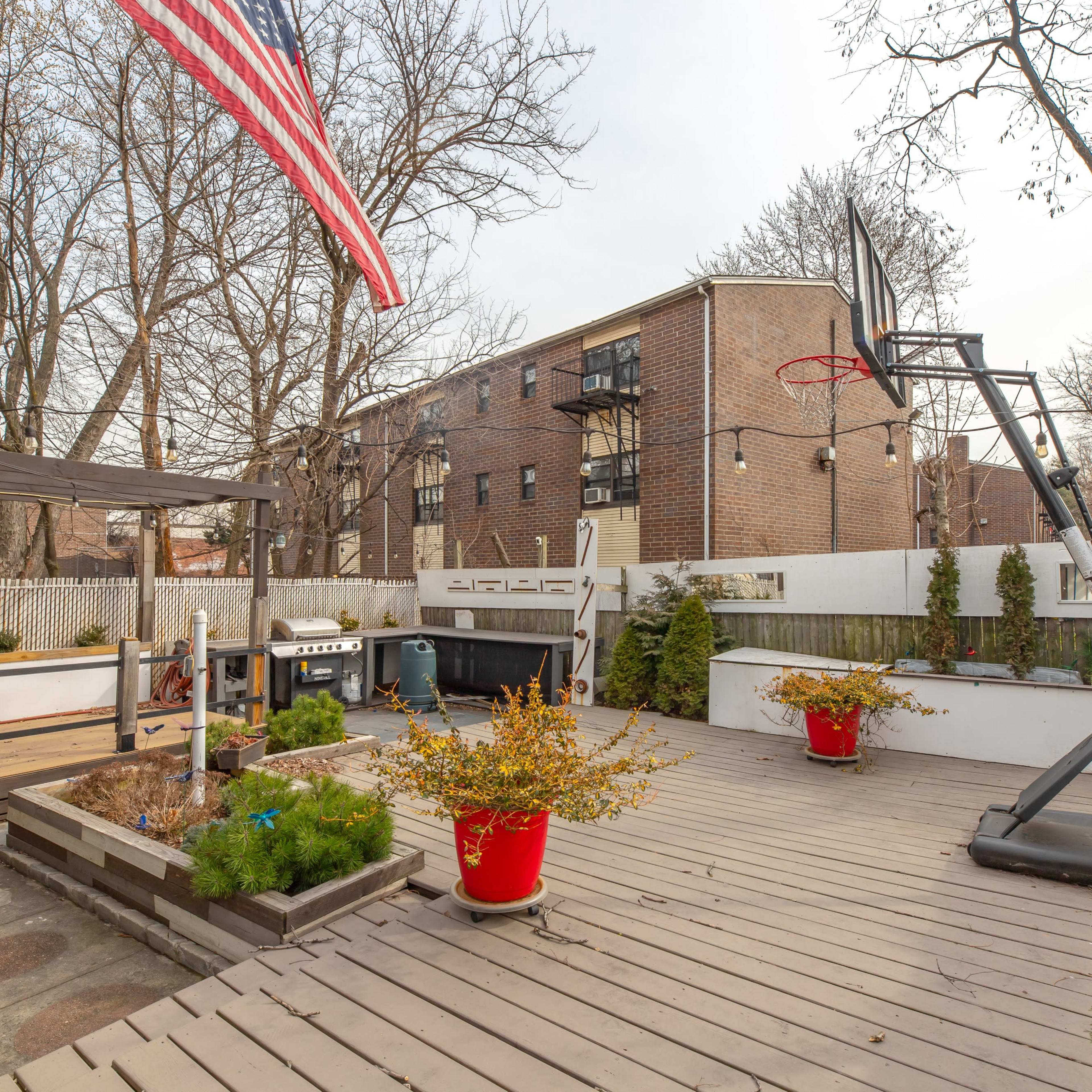 A backyard featuring a wooden deck with potted plants, a basketball hoop, a grill area, and an American flag in the foreground, with an apartment building visible in the background.