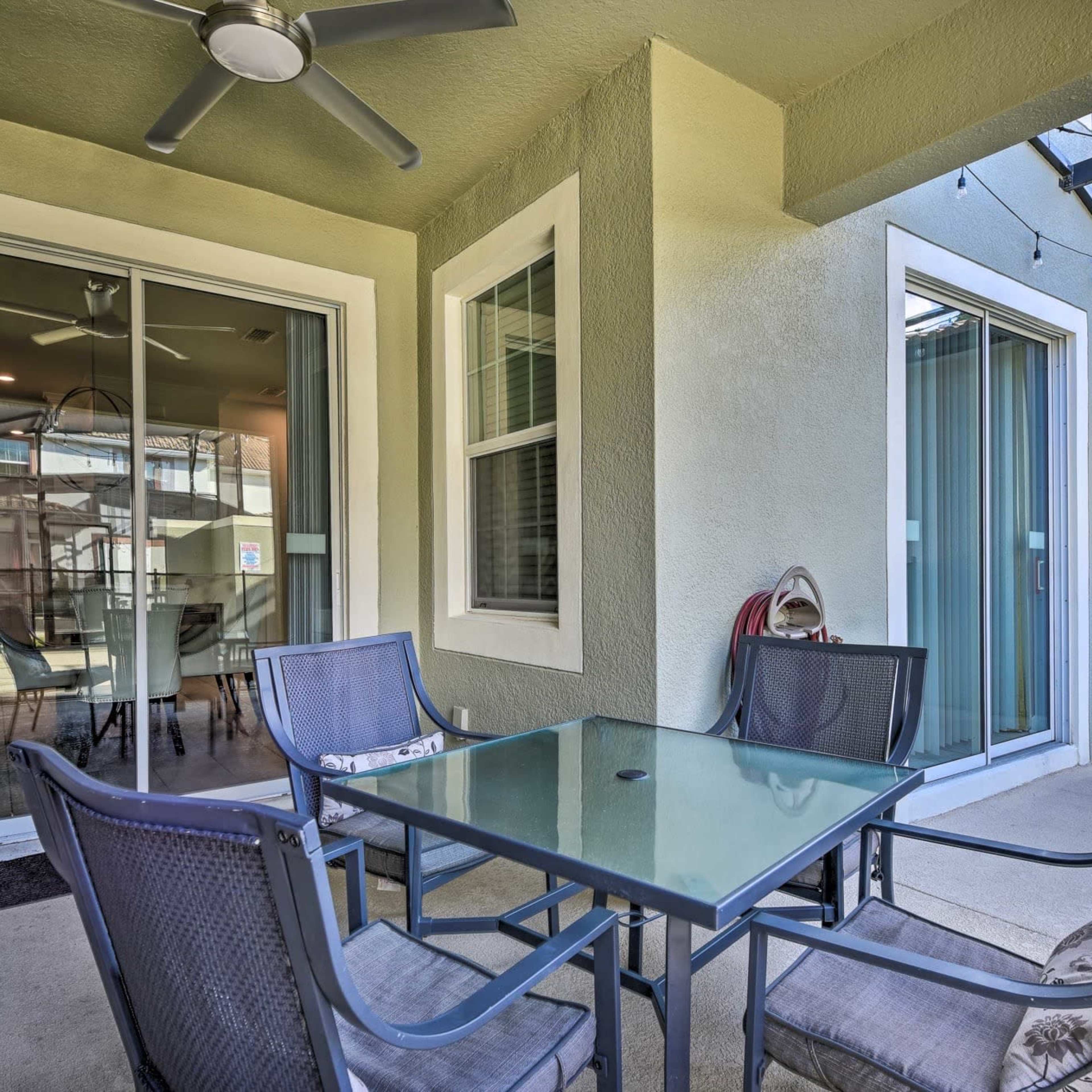 A patio area with a glass table surrounded by four chairs, adjacent to sliding glass doors.