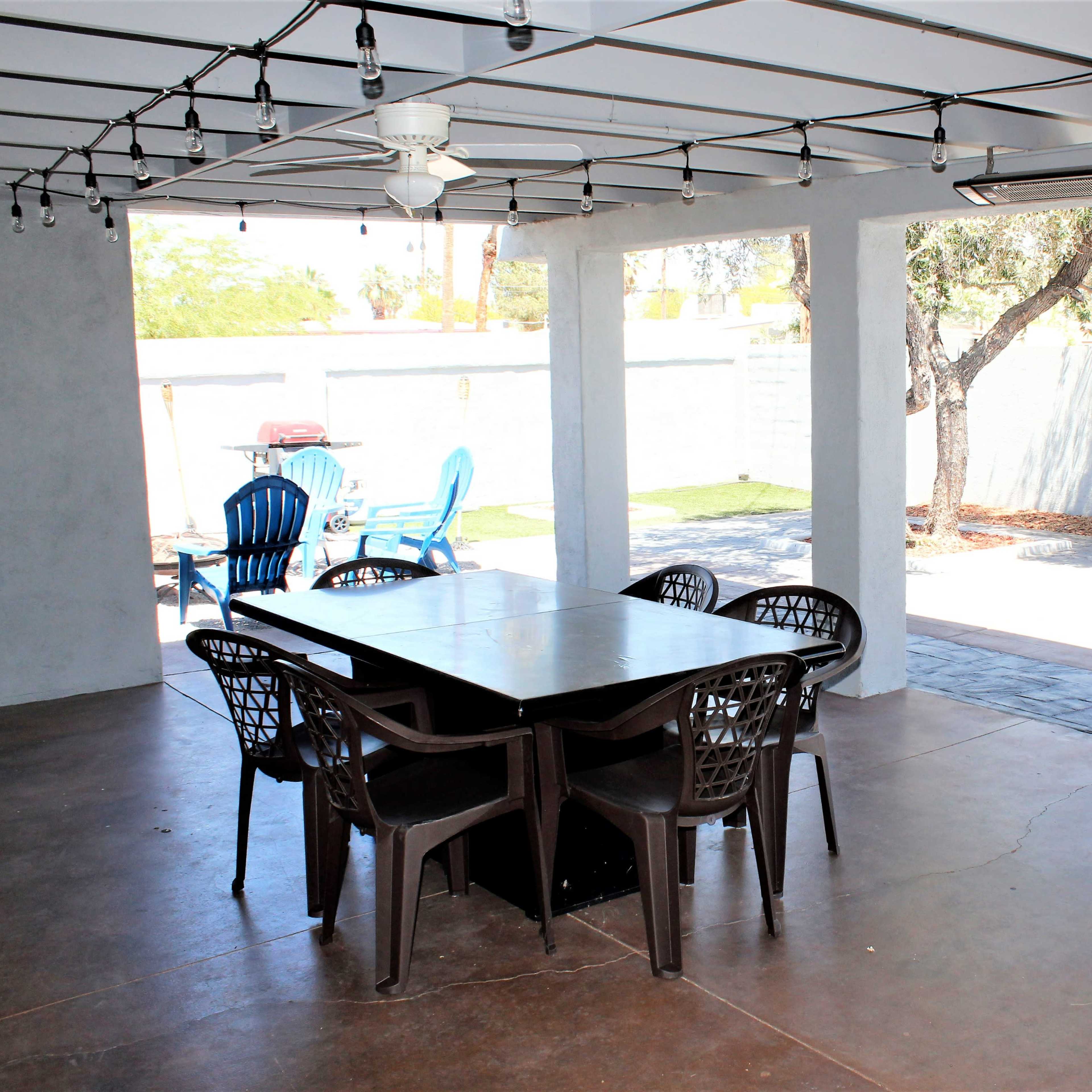 An outdoor patio area with a large table surrounded by chairs, string lights overhead, and additional seating under a shaded space.