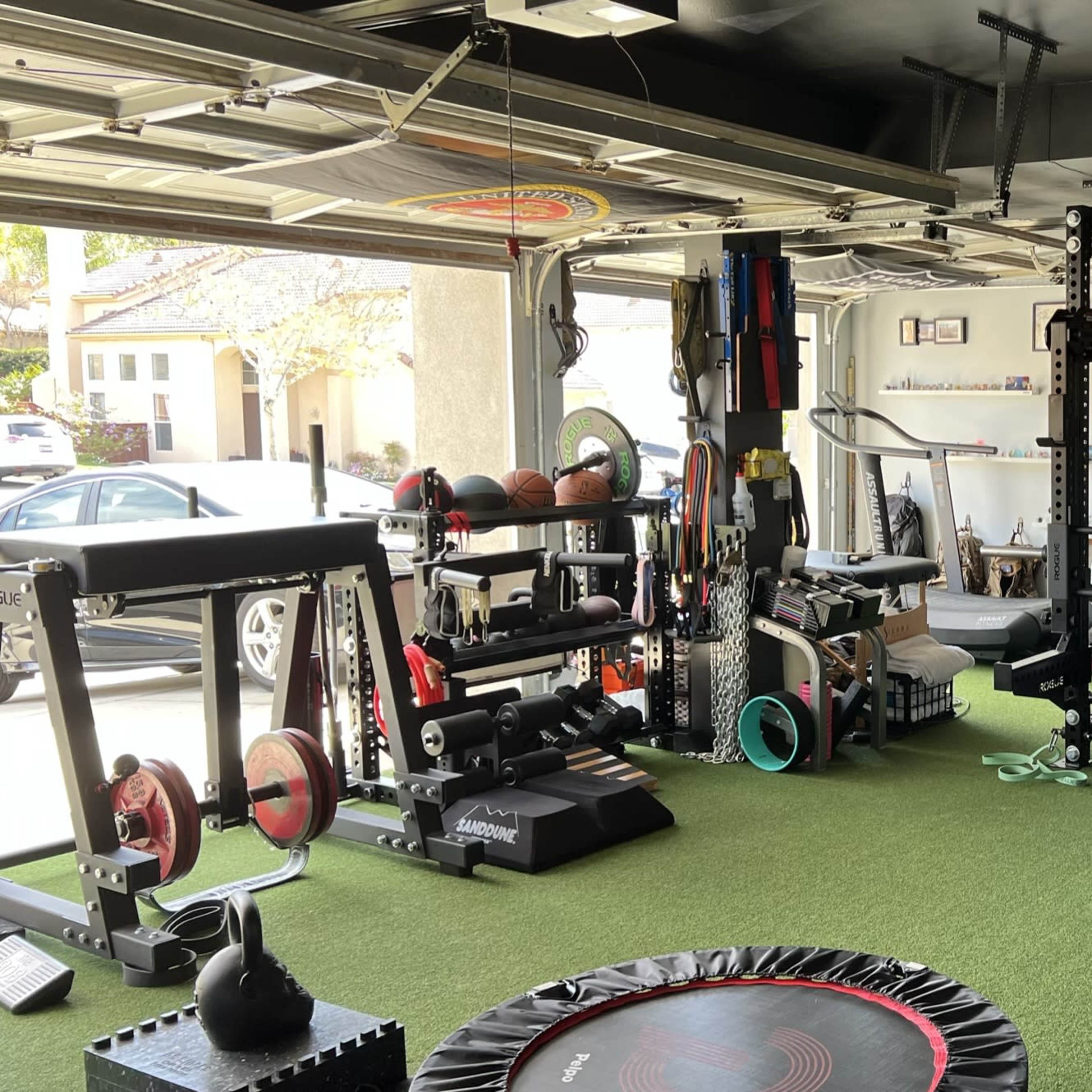 A home garage gym equipped with various fitness machines, weights, and exercise accessories on a green turf floor.