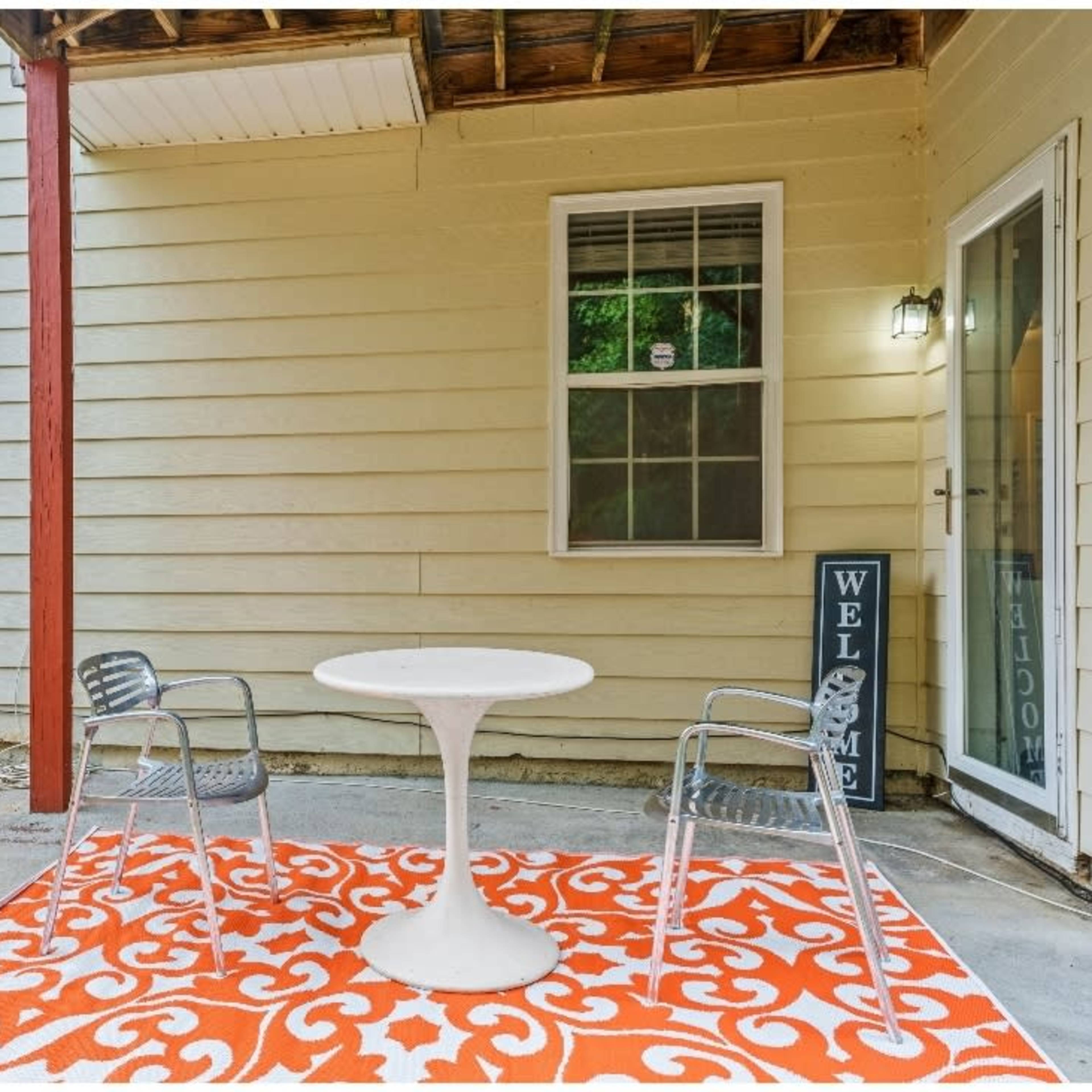 A small patio area with a round white table, two transparent chairs, and an orange patterned rug, surrounded by beige walls and a red wooden fence.