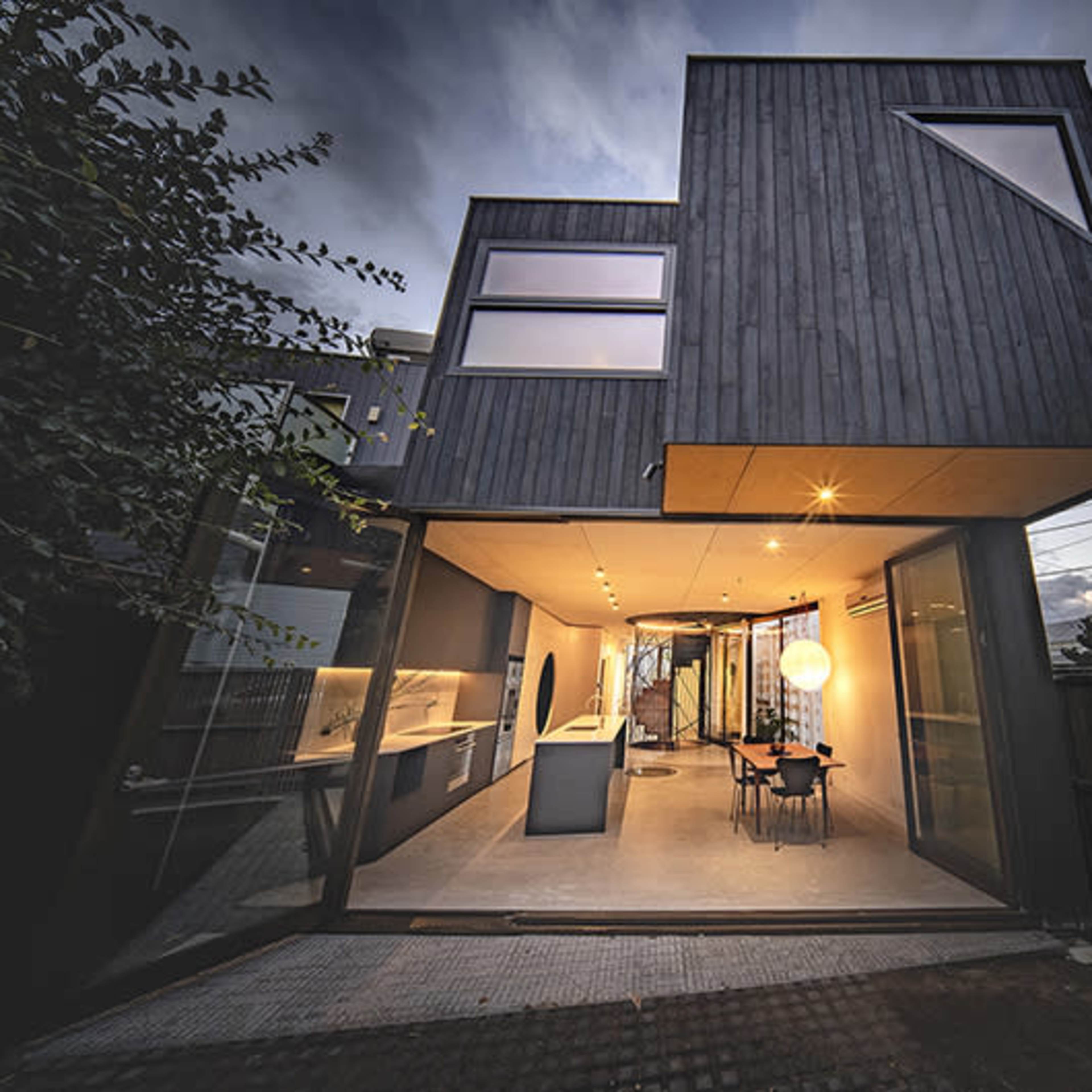 A modern, angular house with a dark wood exterior is shown against a twilight sky, featuring large windows and a spacious open interior connected to an outdoor patio.