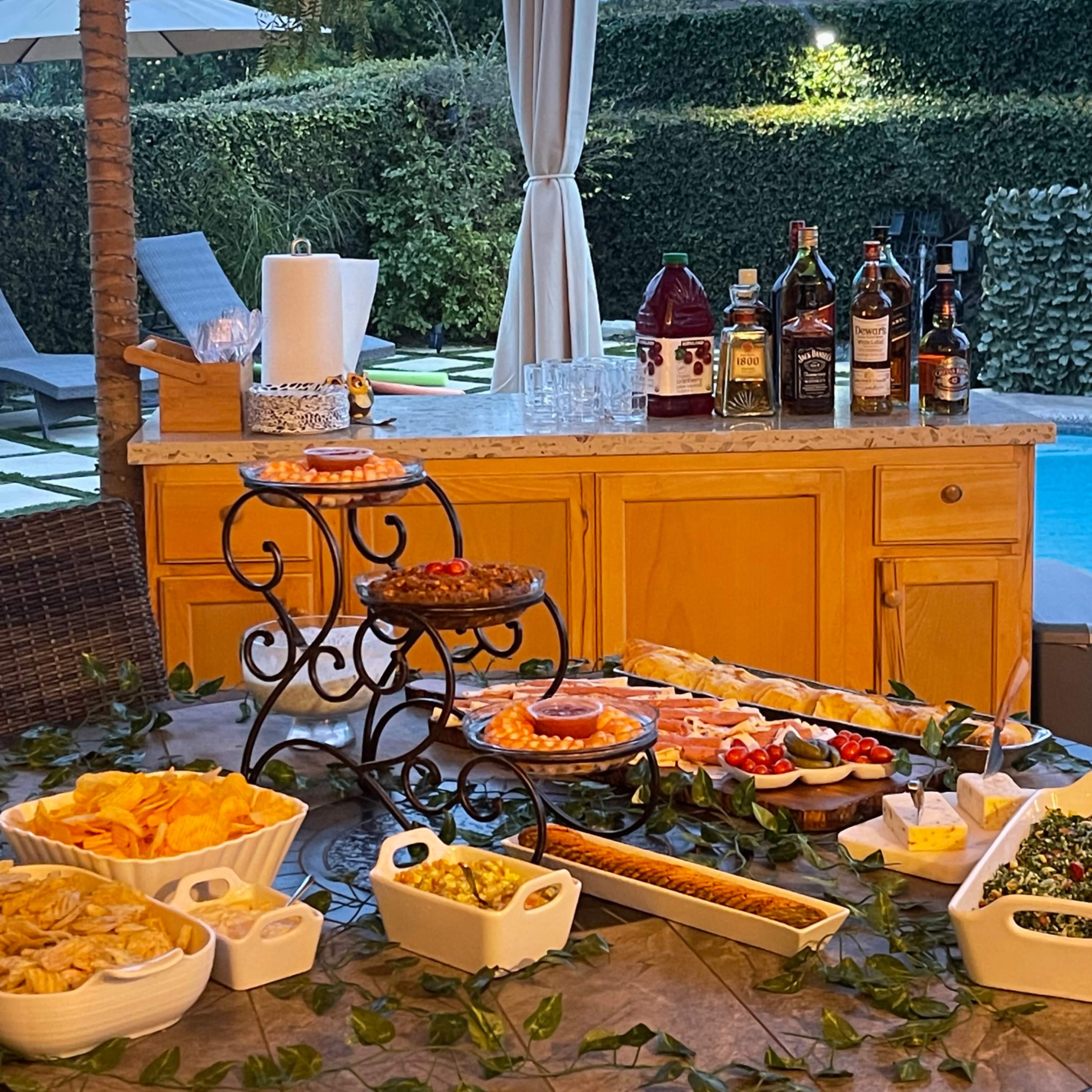 A buffet table is set up outside, featuring various appetizers and drinks, with a swimming pool visible in the background.