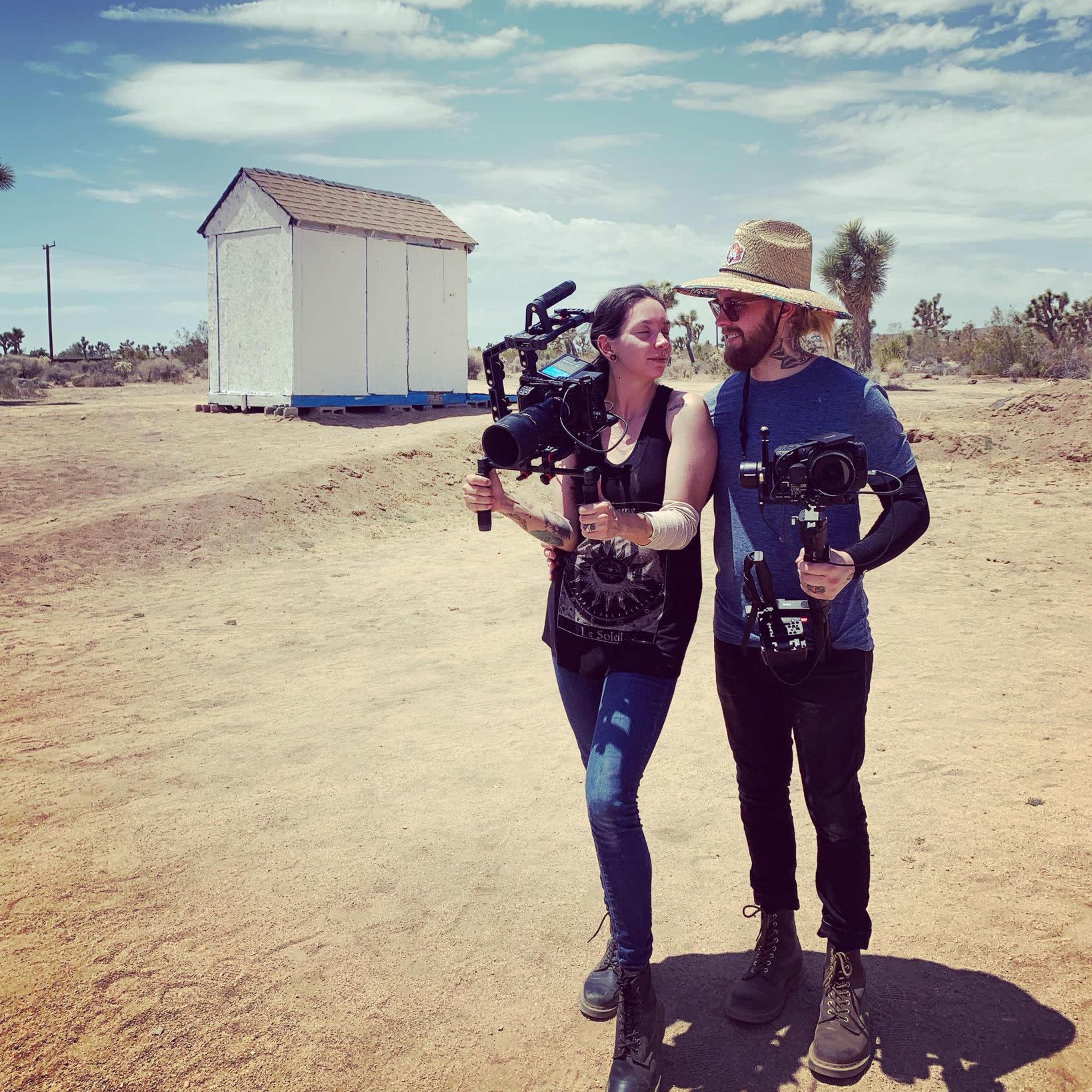 Two individuals hold video cameras while standing in a desert landscape near a small white shed.