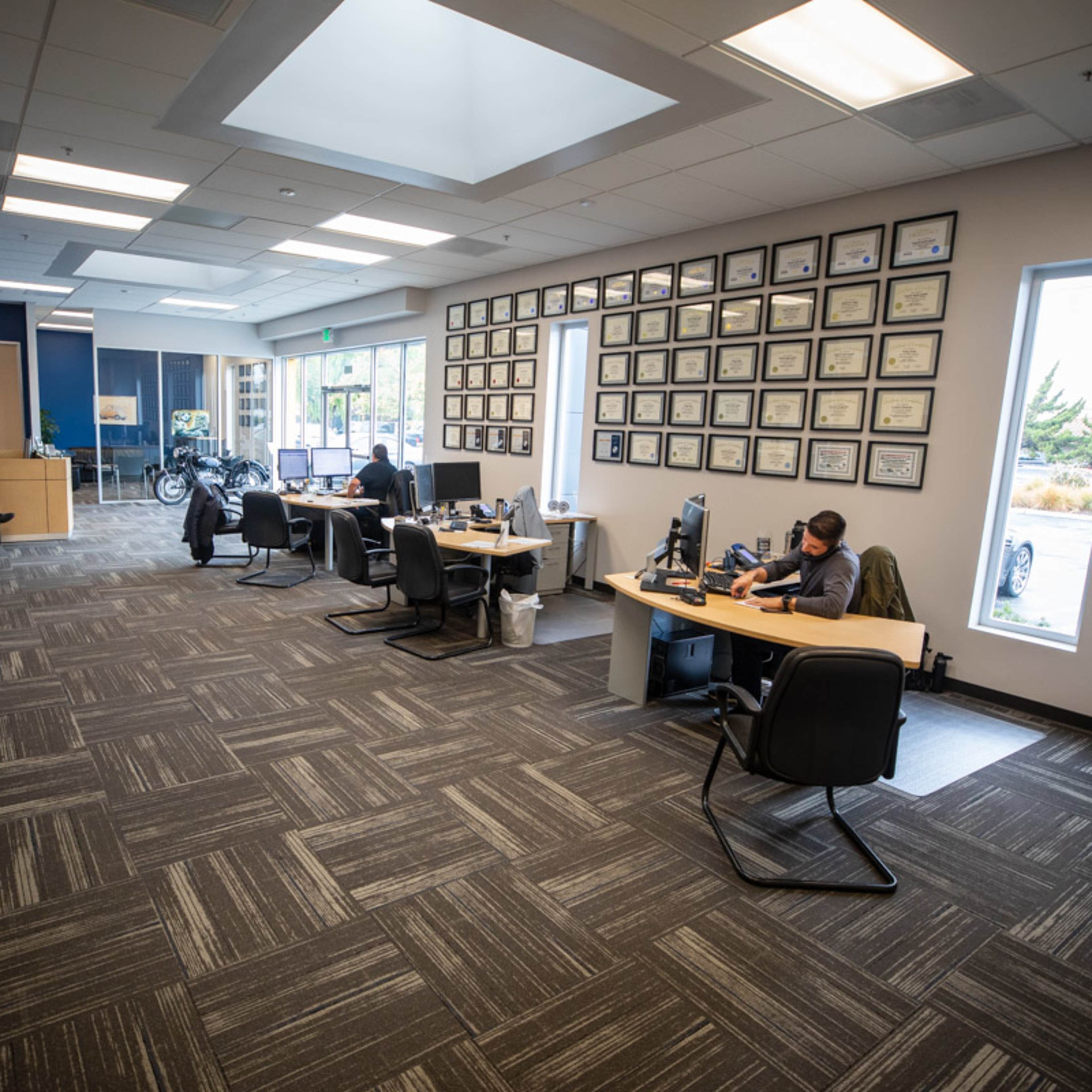 An office space with multiple workstations, a wall adorned with framed certificates, and people working at desks.