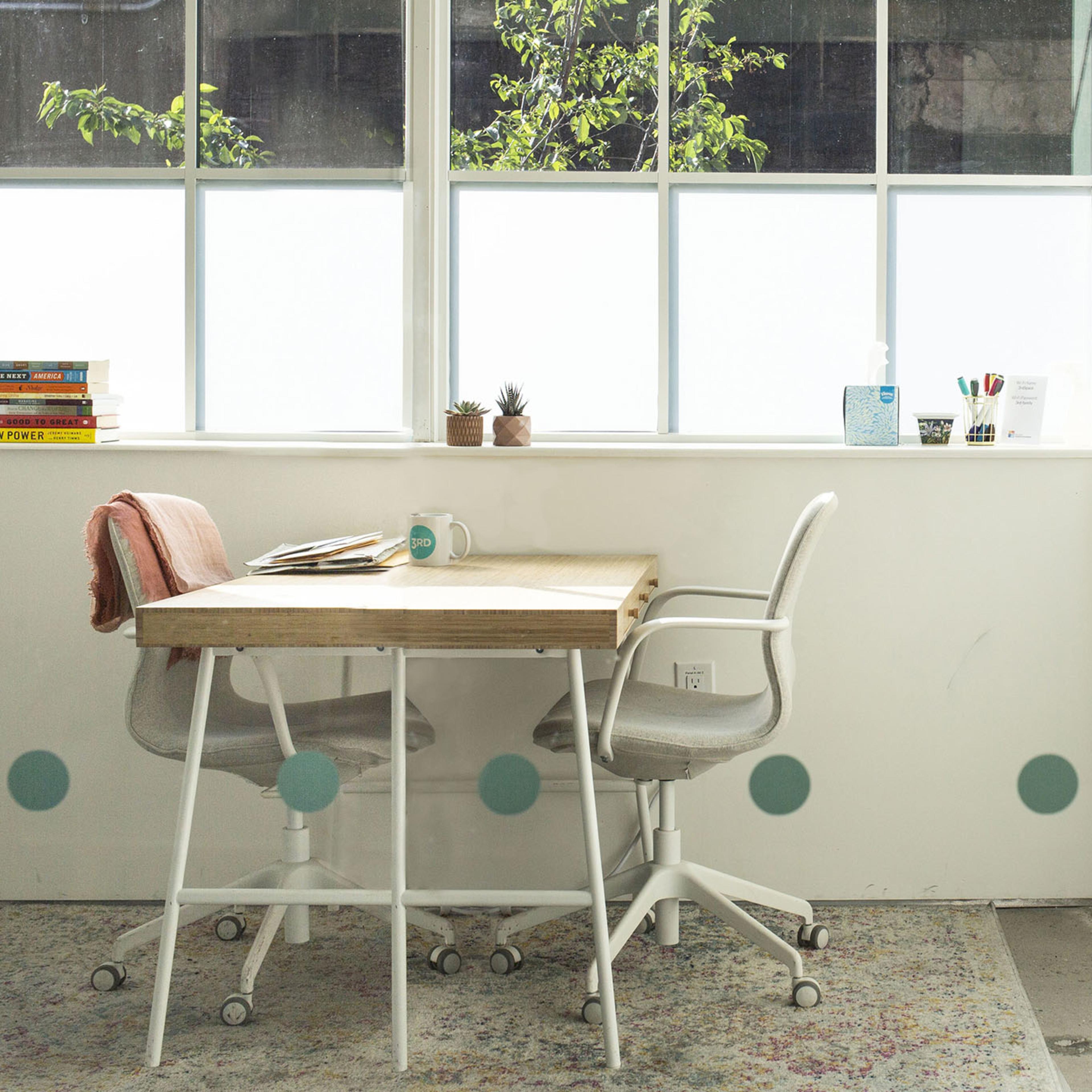 A bright office space featuring a wooden table with two chairs, a window with plants outside, and a printer against the wall.