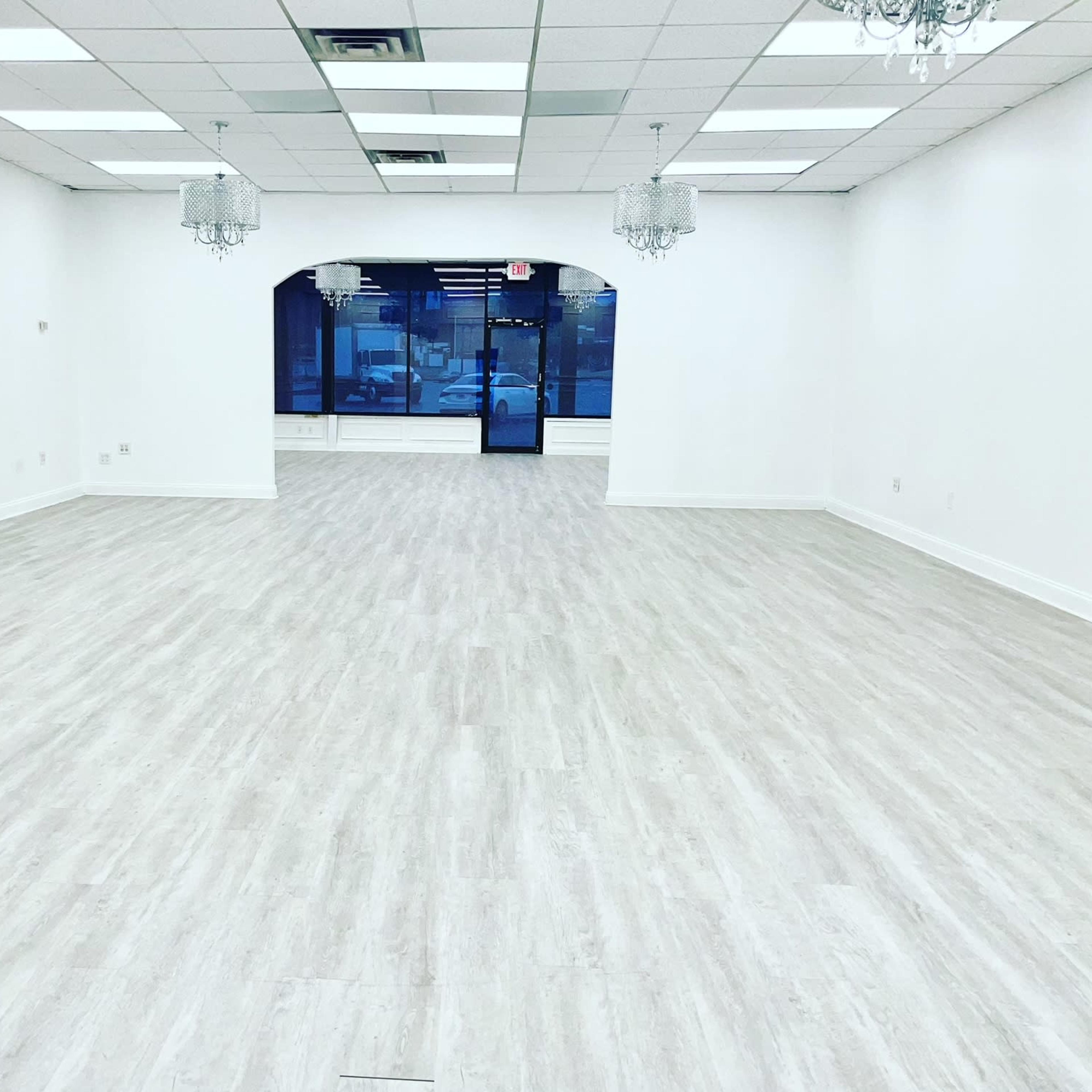 A spacious, empty room with light-colored flooring, white walls, and two crystal chandeliers hanging from the ceiling.
