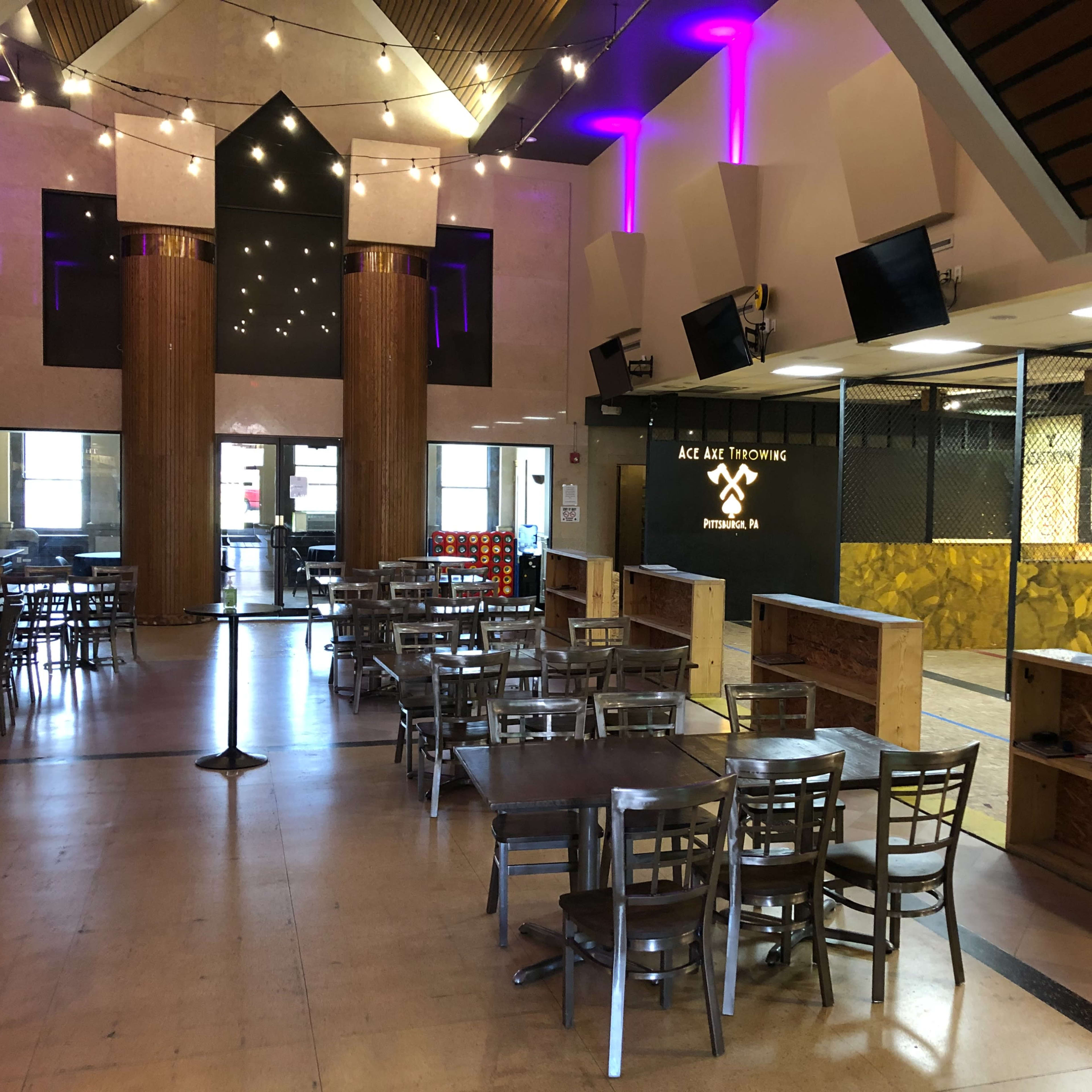 The interior of a venue with rows of metal tables and chairs, a reception area, and a space designed for activities, illuminated with purple lighting.
