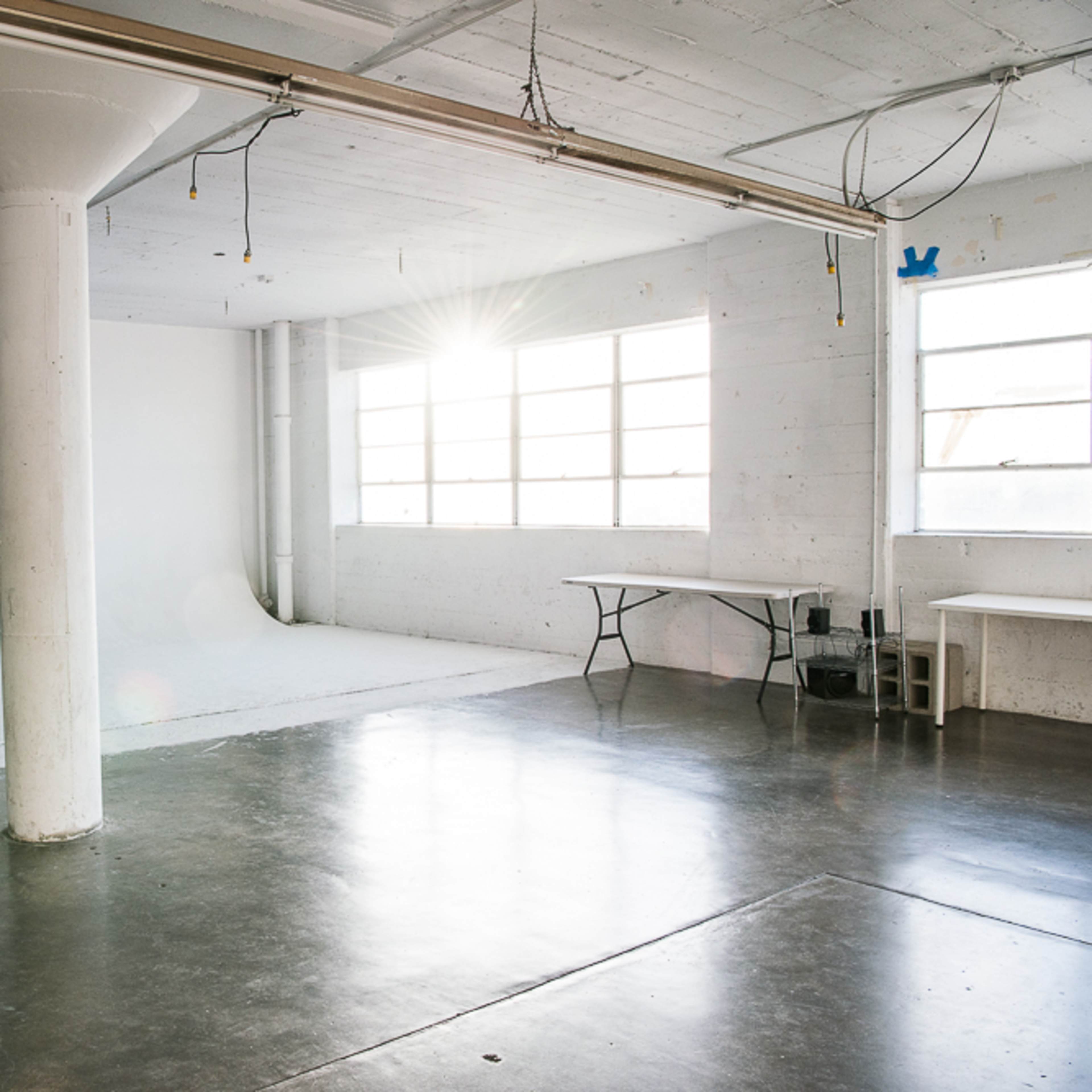 A spacious, empty room with polished concrete floors, large windows, a white backdrop, a black couch, and a table.
