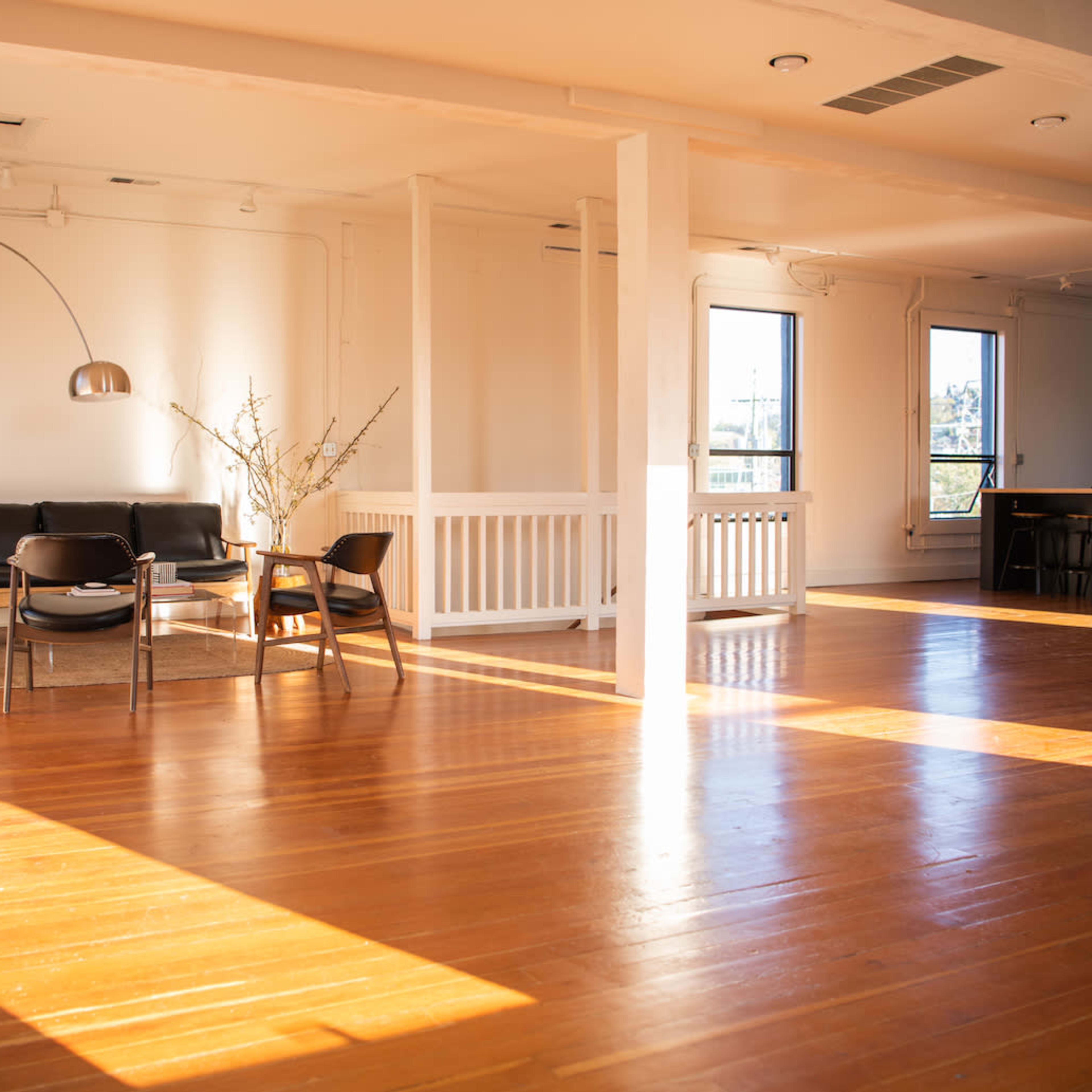 A spacious, well-lit room with polished wooden floors, featuring a black sofa, a large plant, a round pouf, and an arc floor lamp.