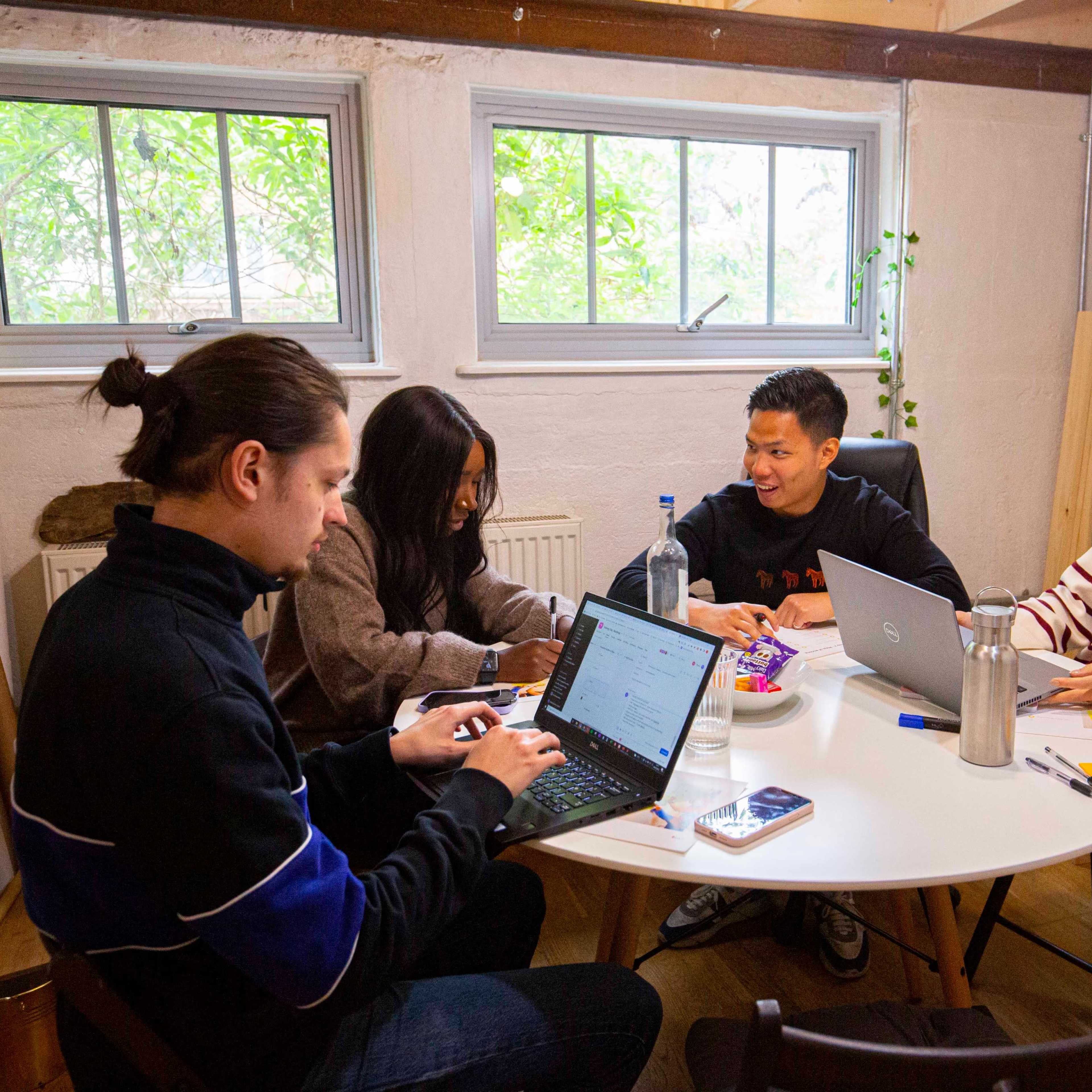 Four individuals sit around a round table in a well-lit room, engaged in discussion while using laptops and snacks are placed on the table.