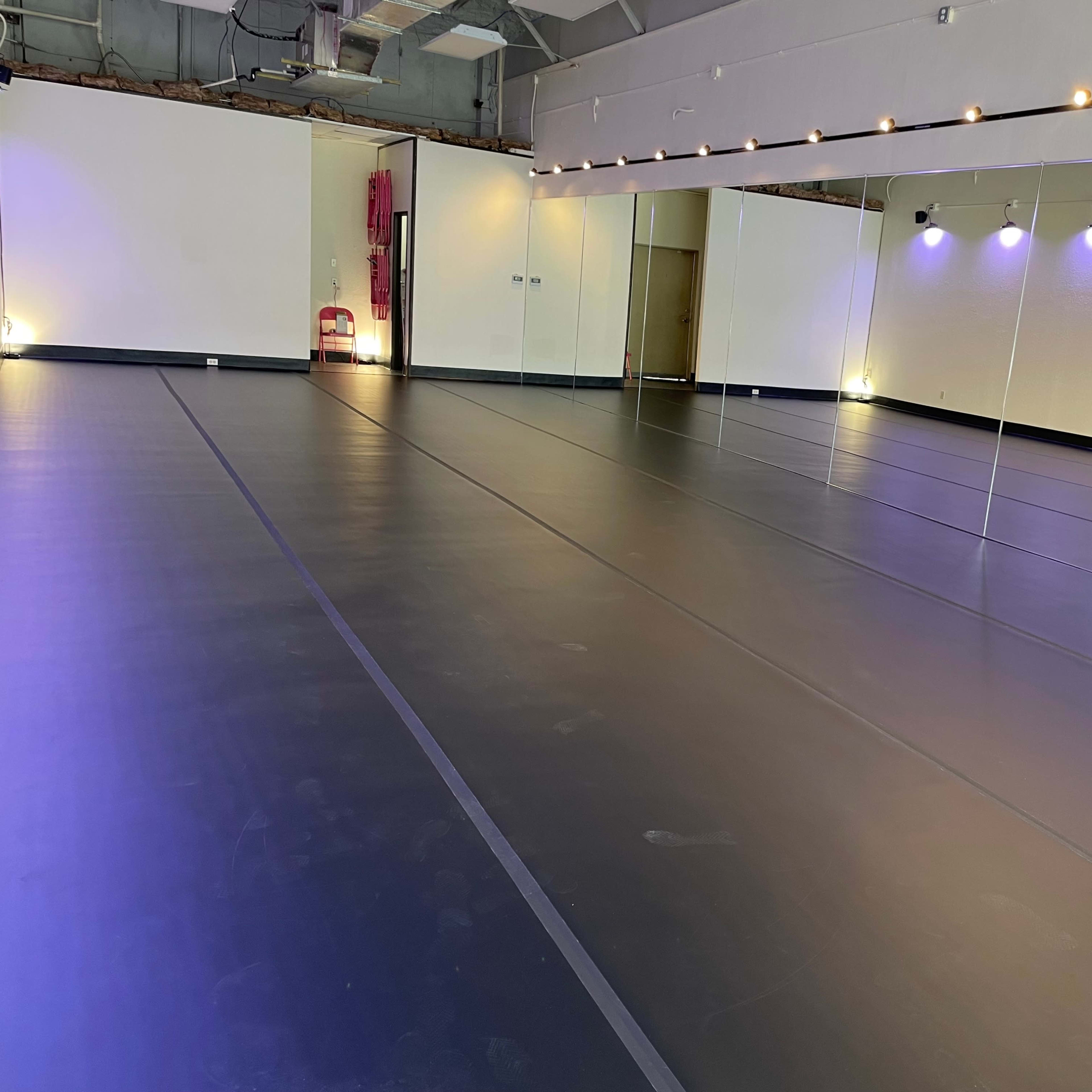 A spacious dance studio with a mirrored wall, black flooring, and soft lighting.