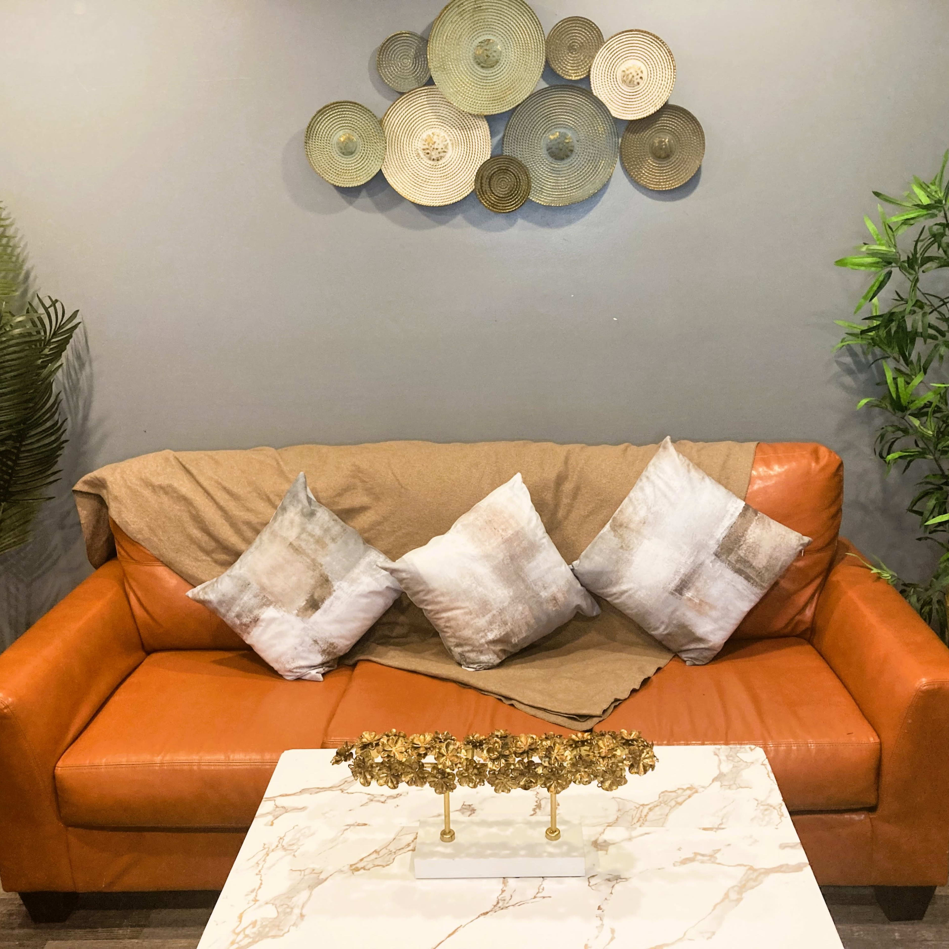A brown leather sofa with decorative pillows and a marble-top coffee table, set against a gray wall decorated with circular wall art and flanked by potted plants.