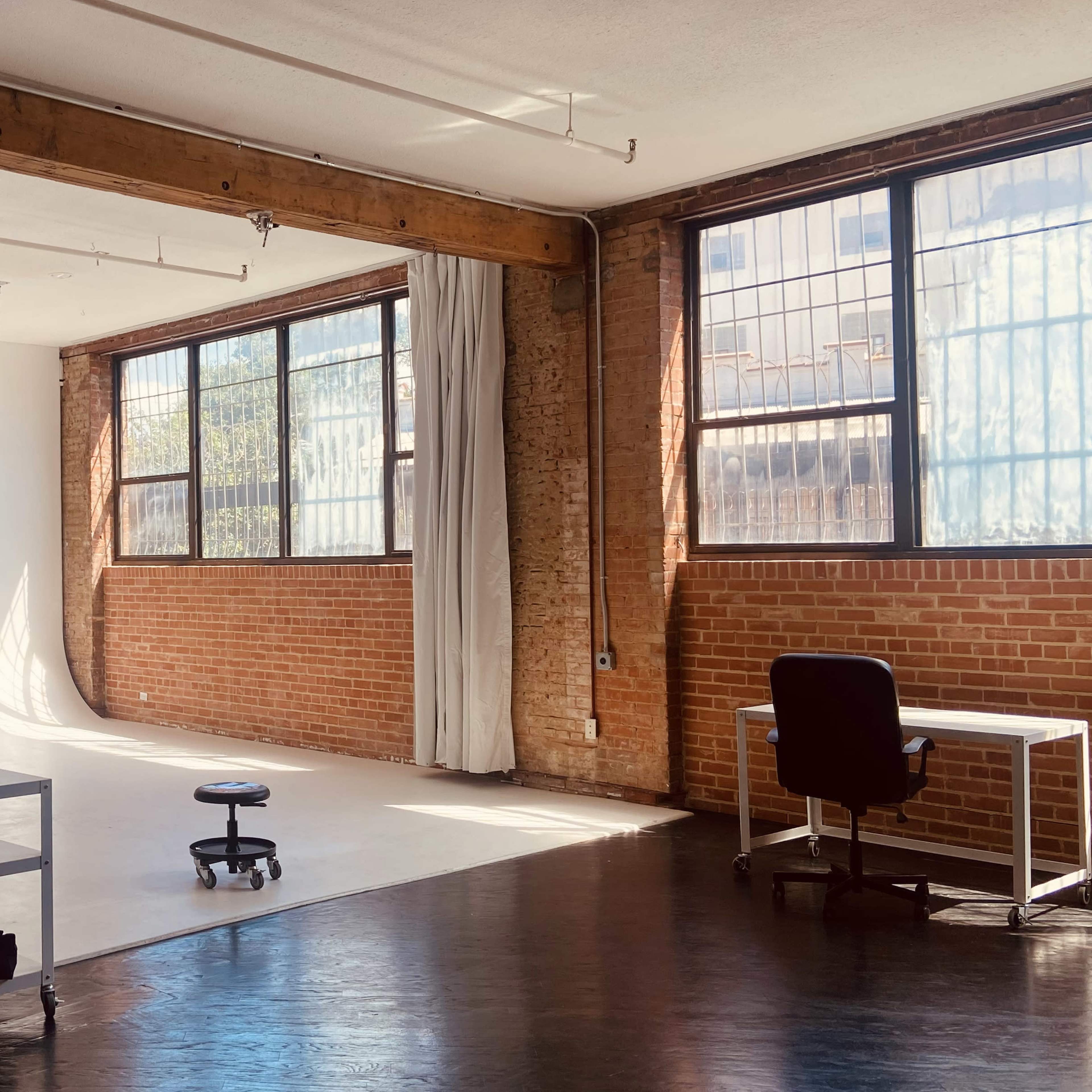 A spacious studio with large windows, brick walls, and a white backdrop in one corner, featuring a rolling stool and a desk.