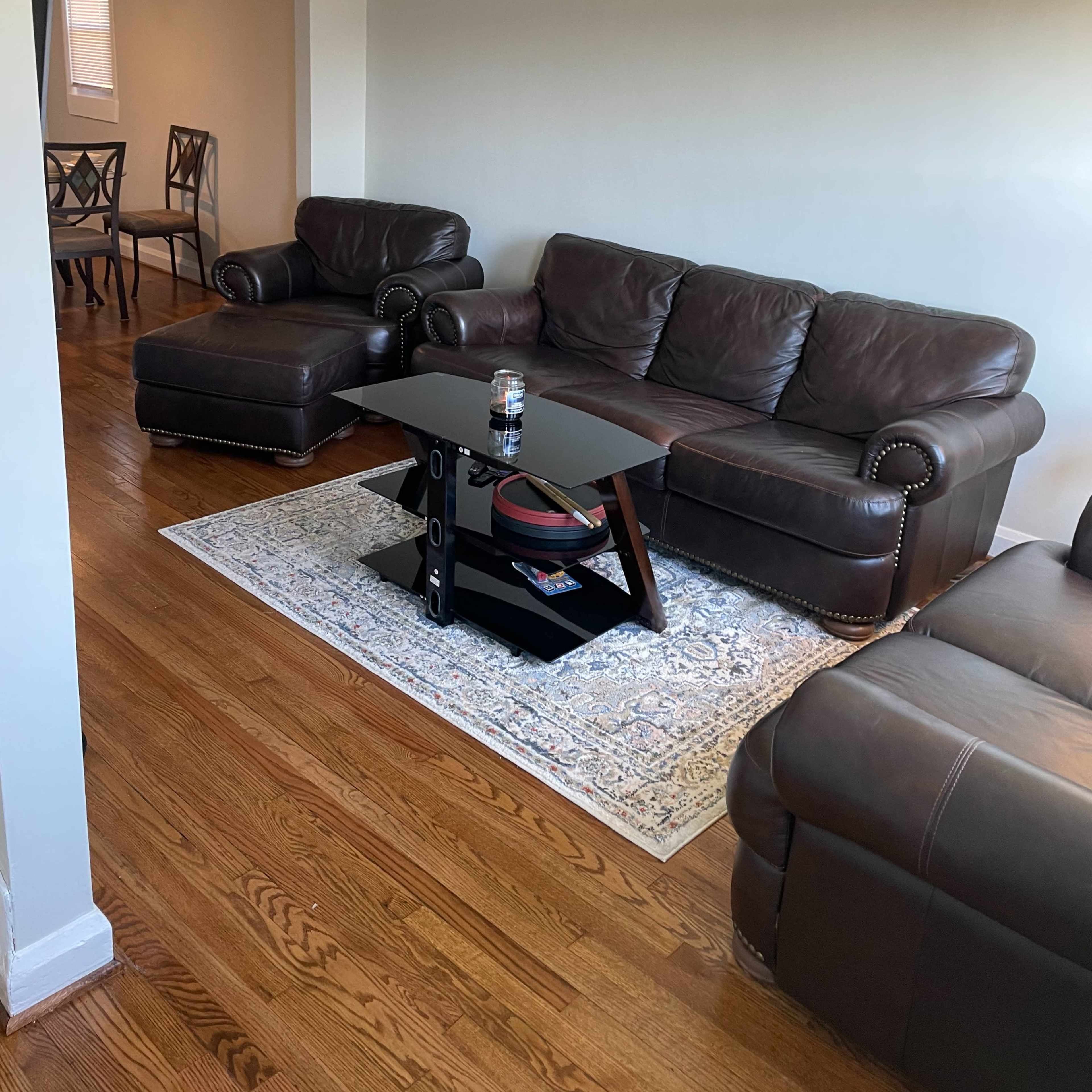 The living room features a brown leather sectional sofa, a glass-top coffee table, and a light-colored area rug on hardwood flooring.