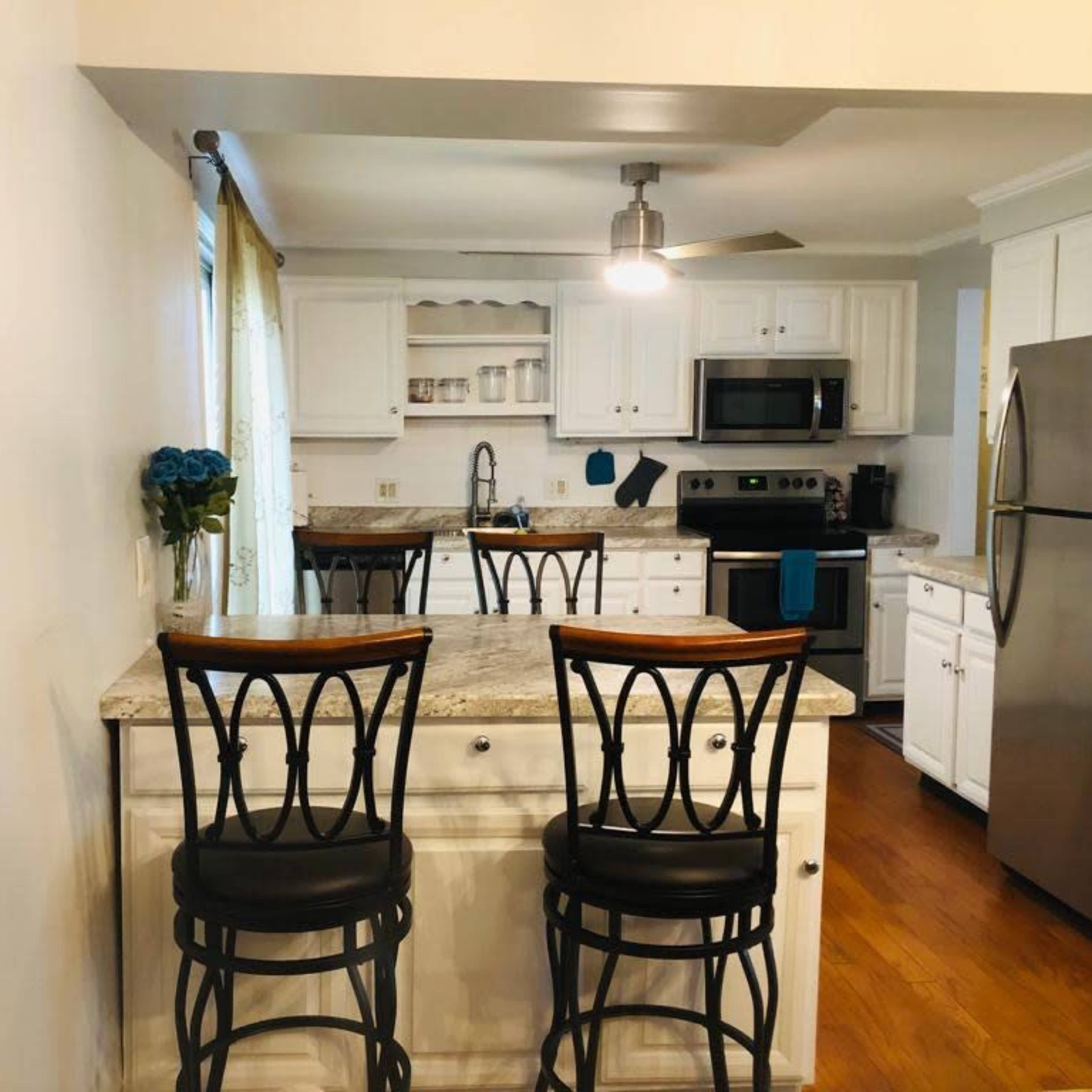 A bright kitchen features white cabinetry, granite countertops with two black bar stools, and modern appliances.