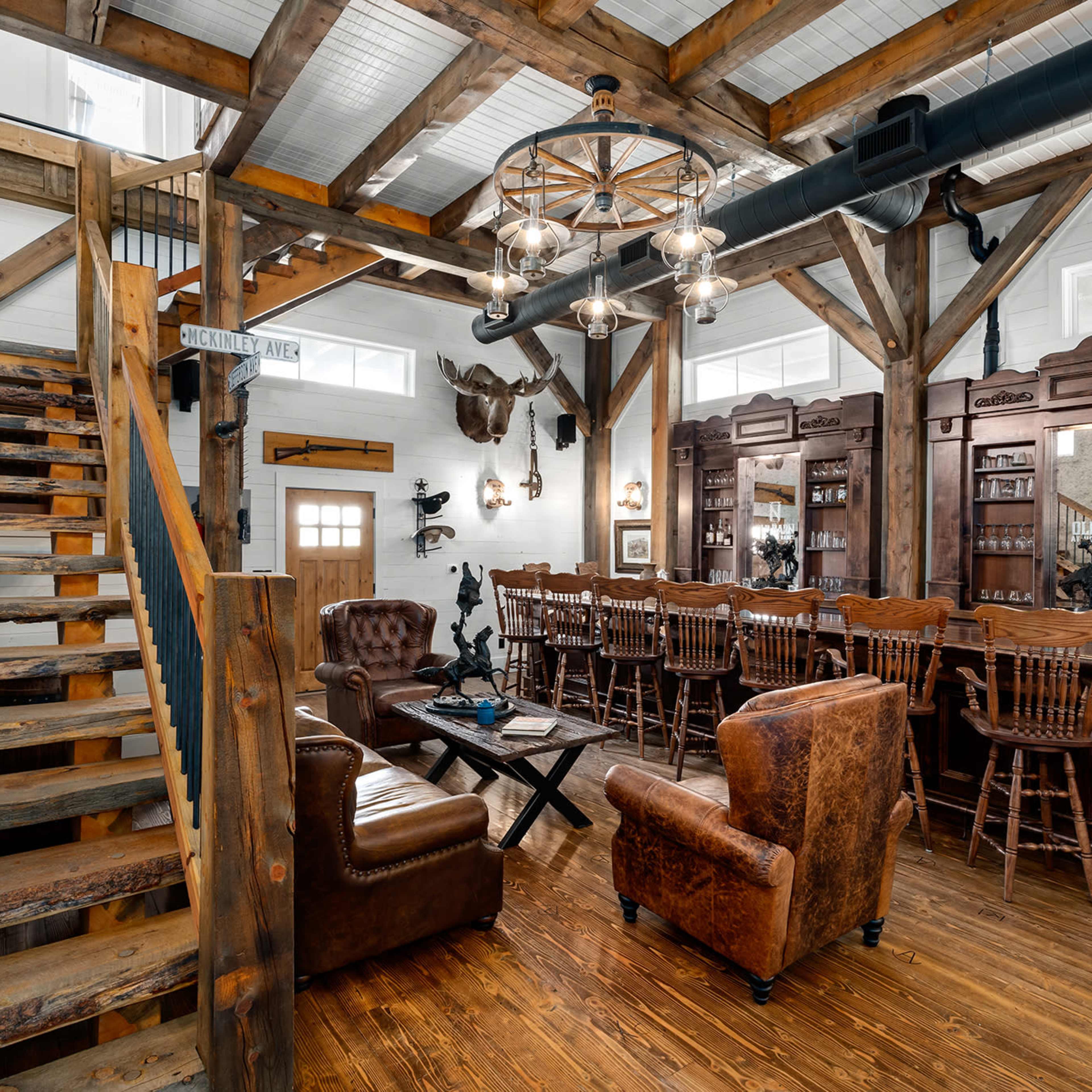 A rustic interior of a bar with wooden beams, a staircase, and a long counter with high stools, complemented by leather seating and decorative elements.