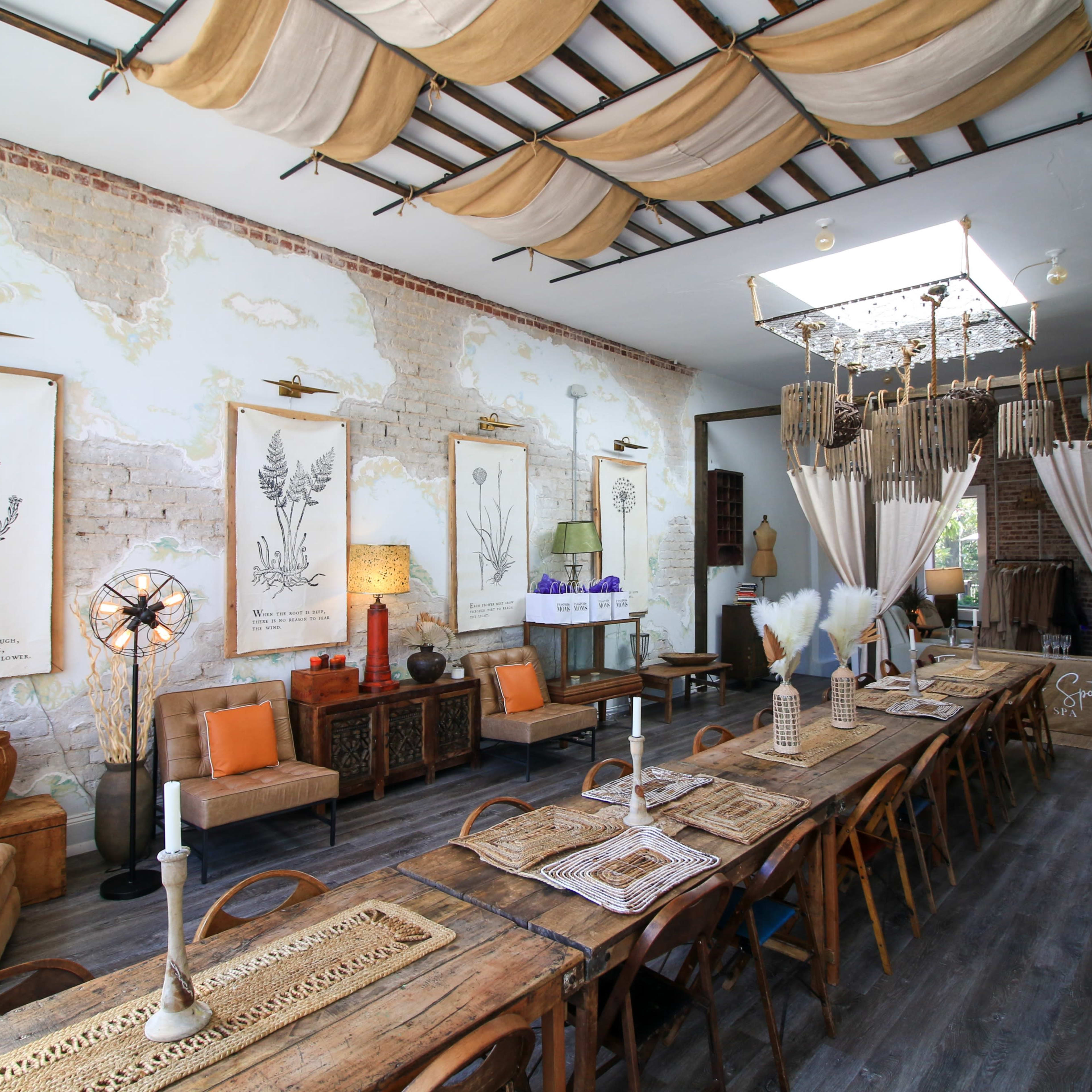 A spacious dining area featuring a long wooden table surrounded by various seating options, with botanical artwork displayed on the walls and soft lighting hanging from the ceiling.