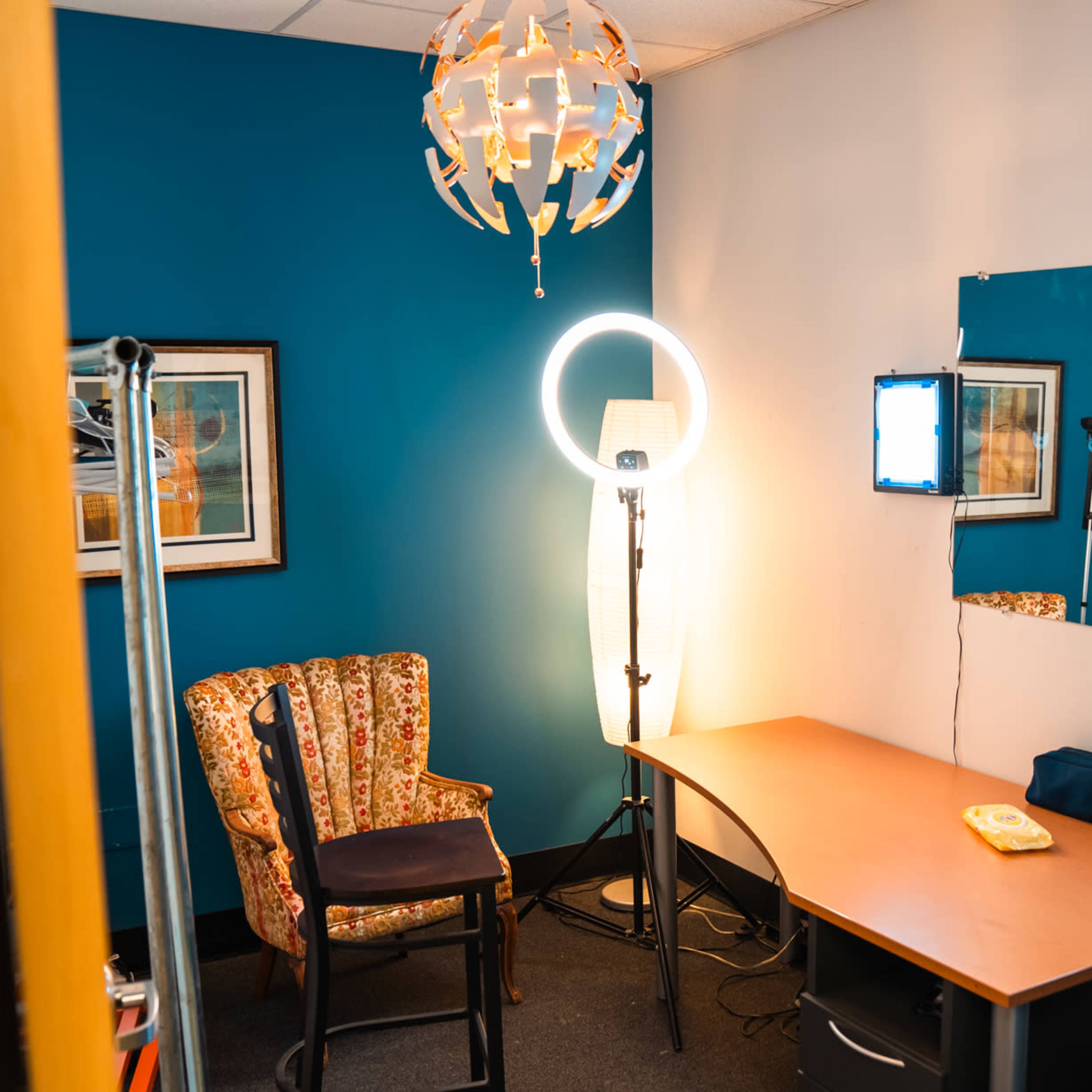 A small makeup room with a colorful chair, a circular light stand, and a desk with a phone.