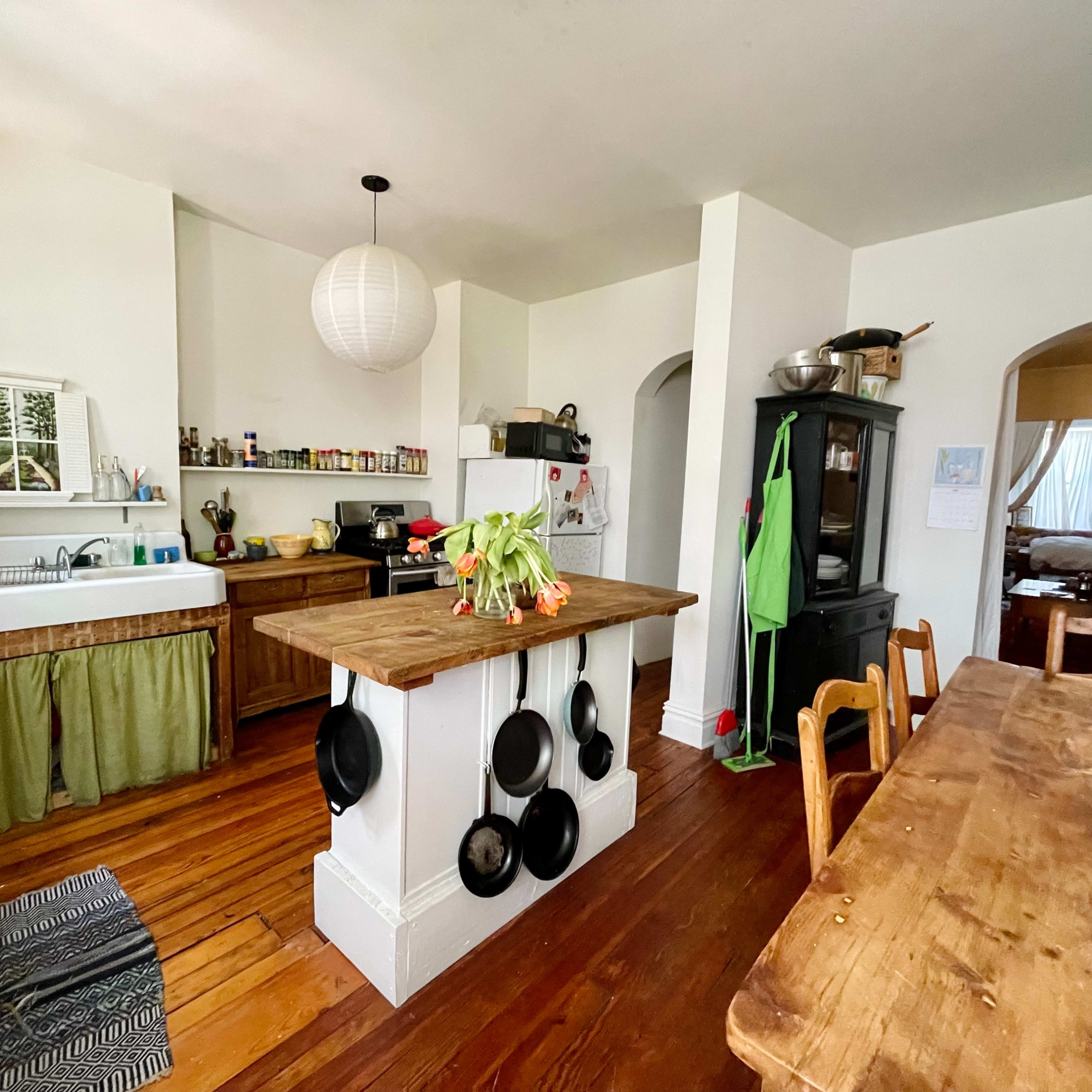 A kitchen features wooden cabinets, an island with hanging pots, and a dining table set with a vase of flowers.