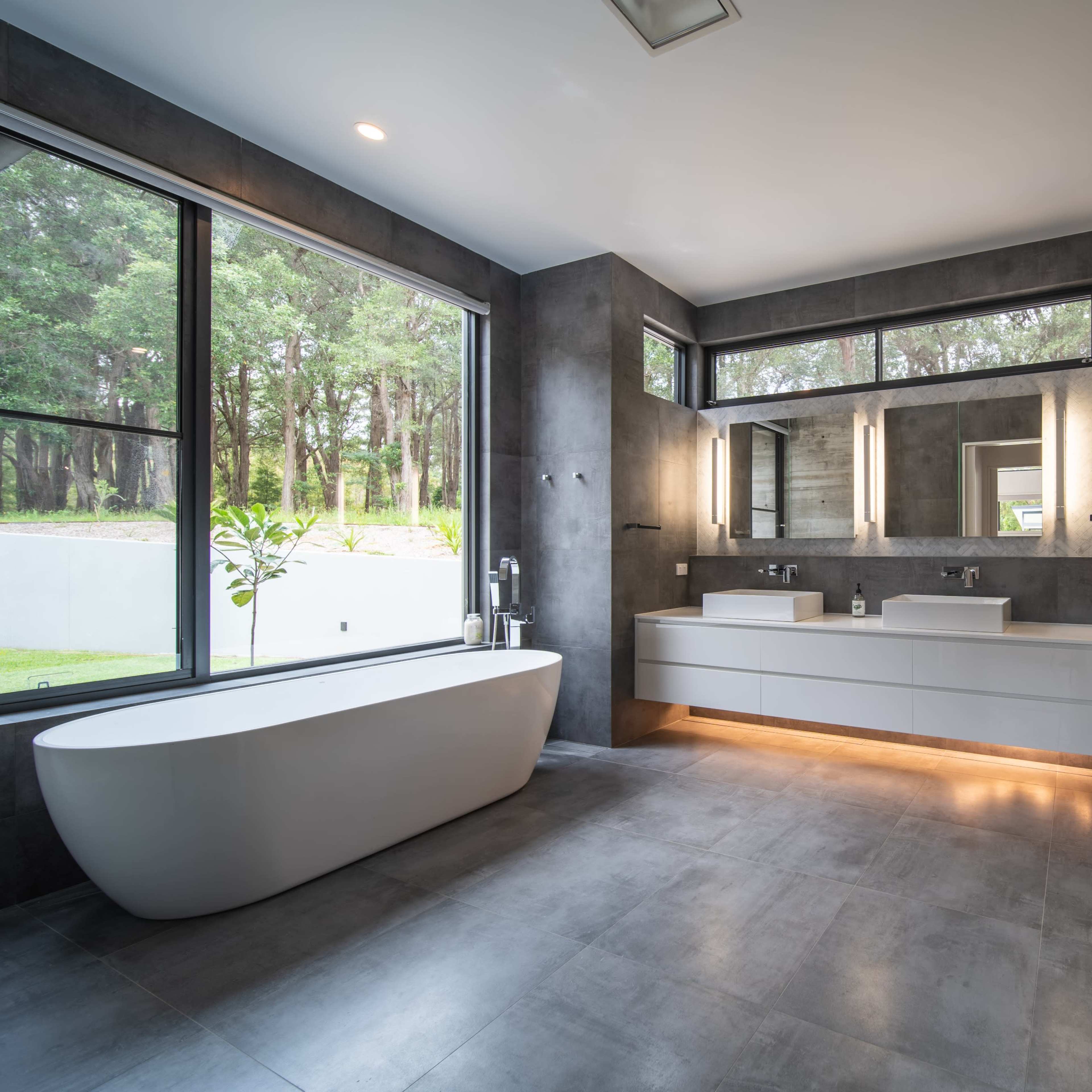 A modern bathroom featuring a freestanding bathtub, dual sinks with ample counter space, and large windows overlooking greenery.