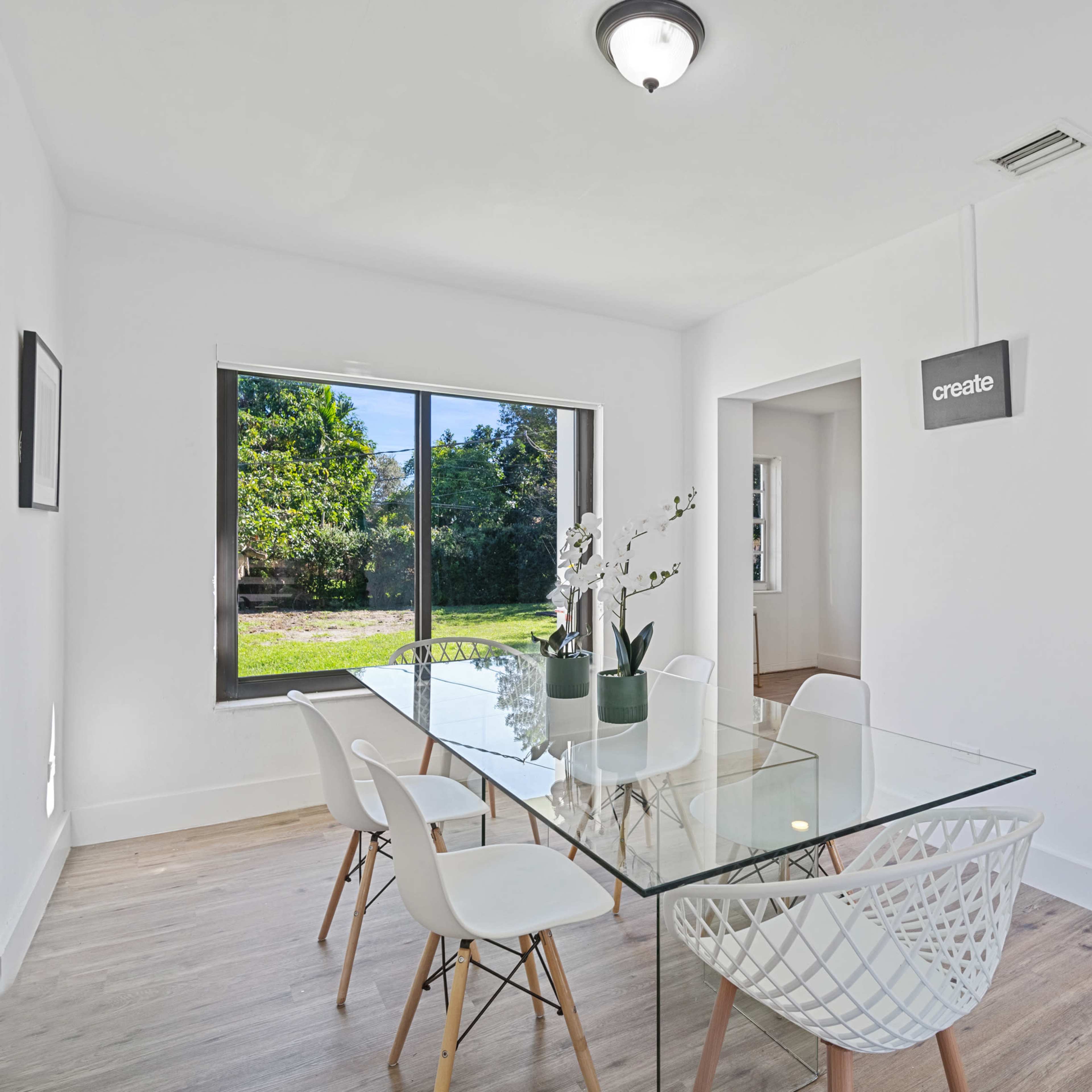 A modern dining area features a glass table surrounded by white chairs, with a view of a green outdoor space through a large window.
