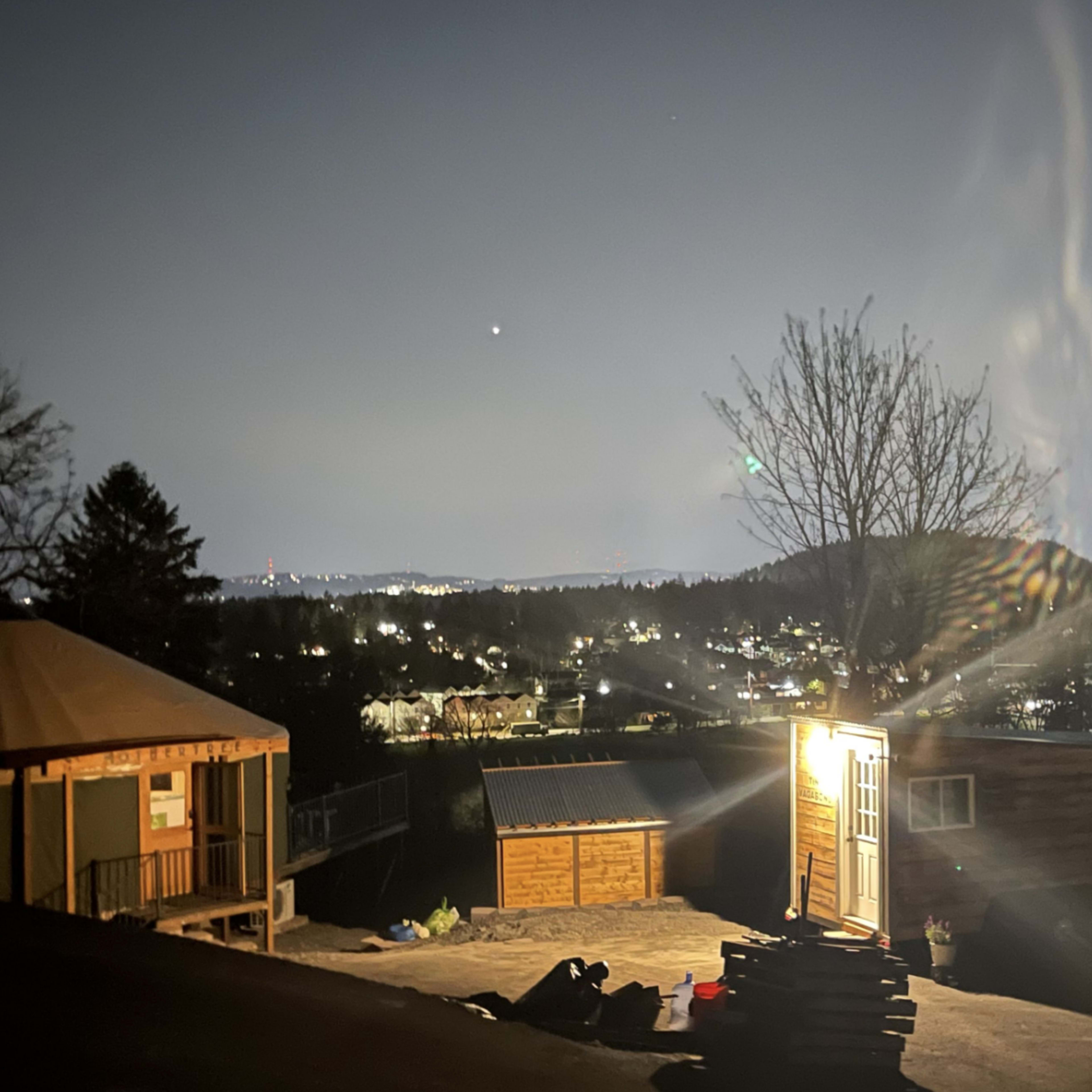 A brightly lit cabin stands on a hillside overlooking a city illuminated by night lights.