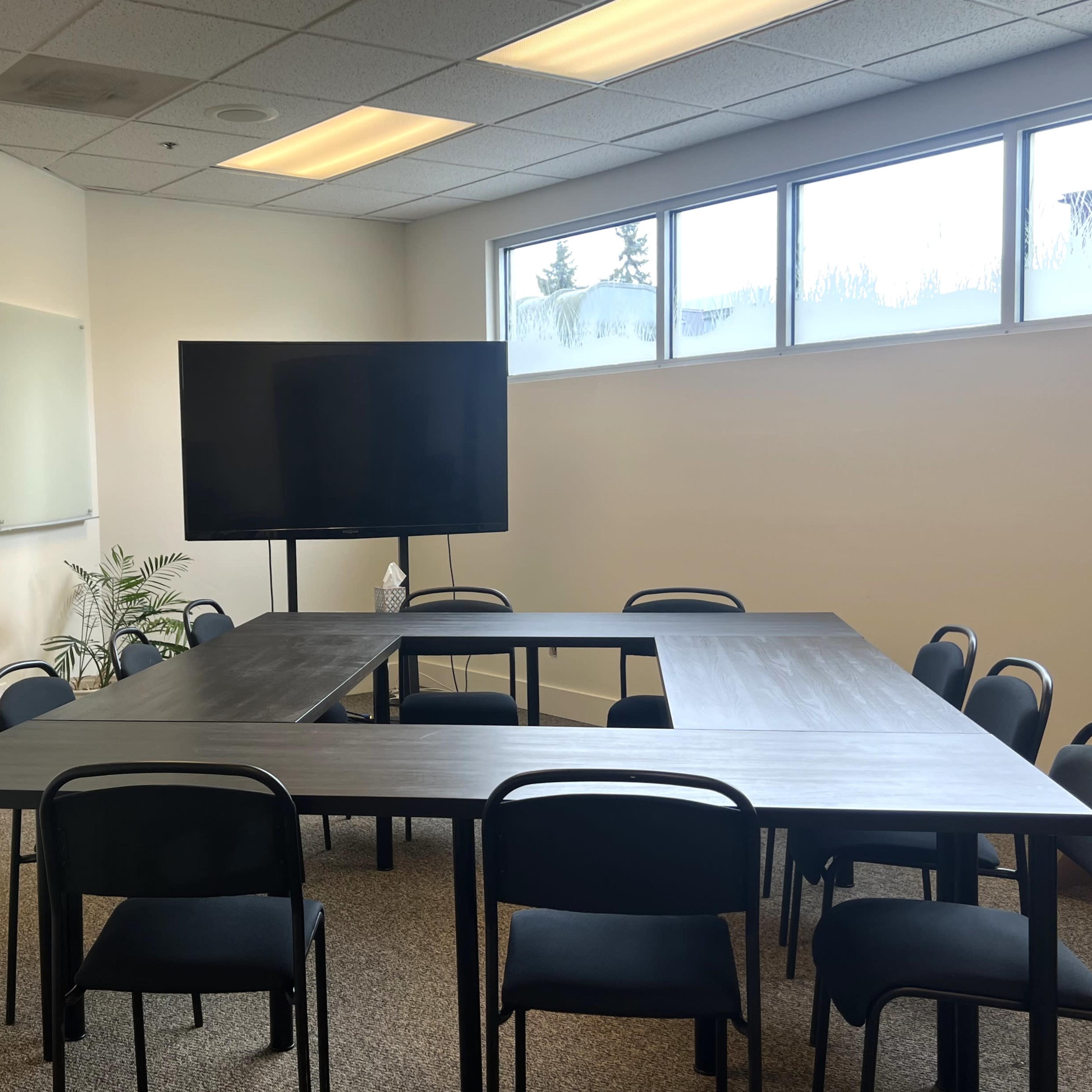 A conference room with a large rectangular table surrounded by black chairs, a television screen on a stand, and whiteboards on the wall.