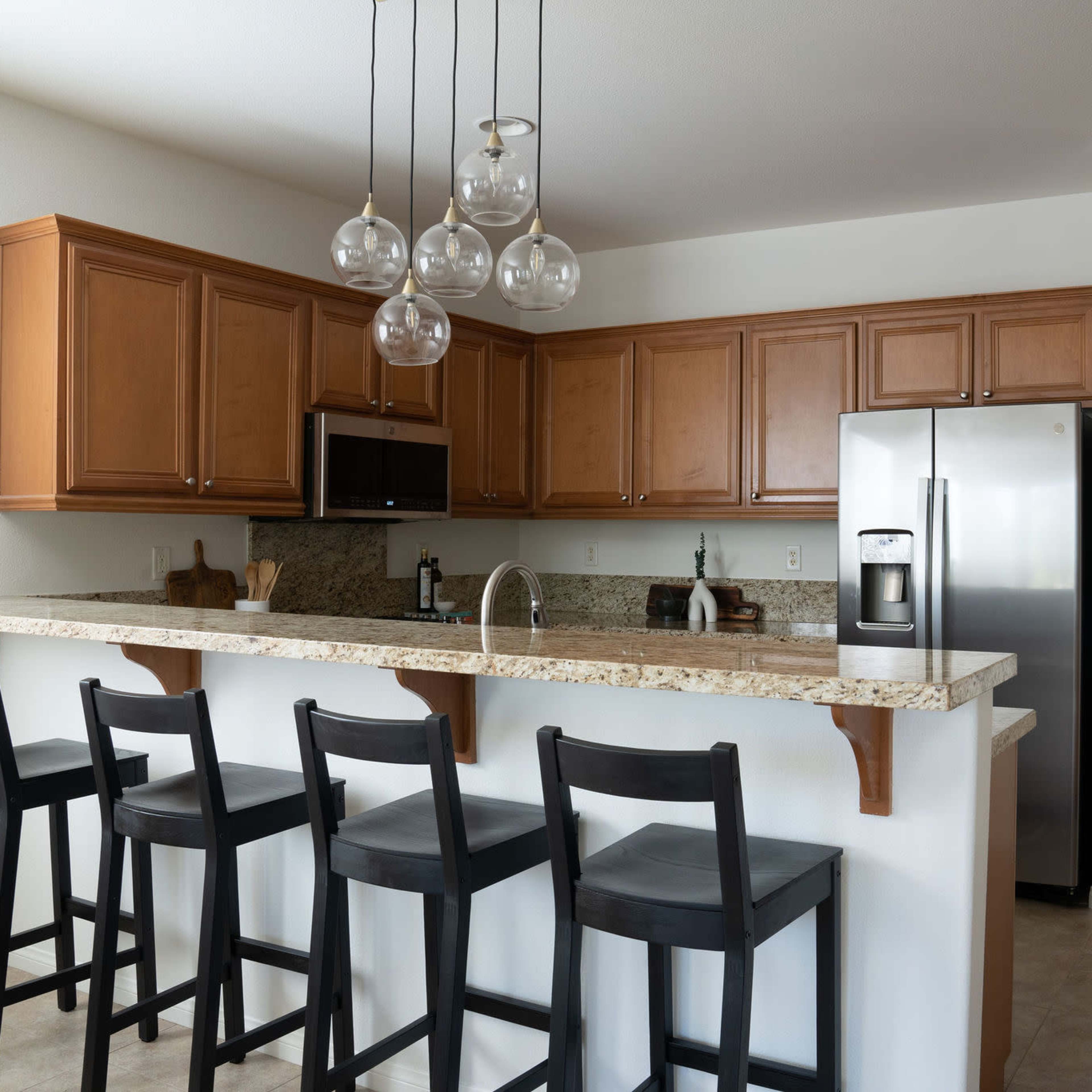 A modern kitchen features wooden cabinets, stainless steel appliances, and a bar with four black stools.