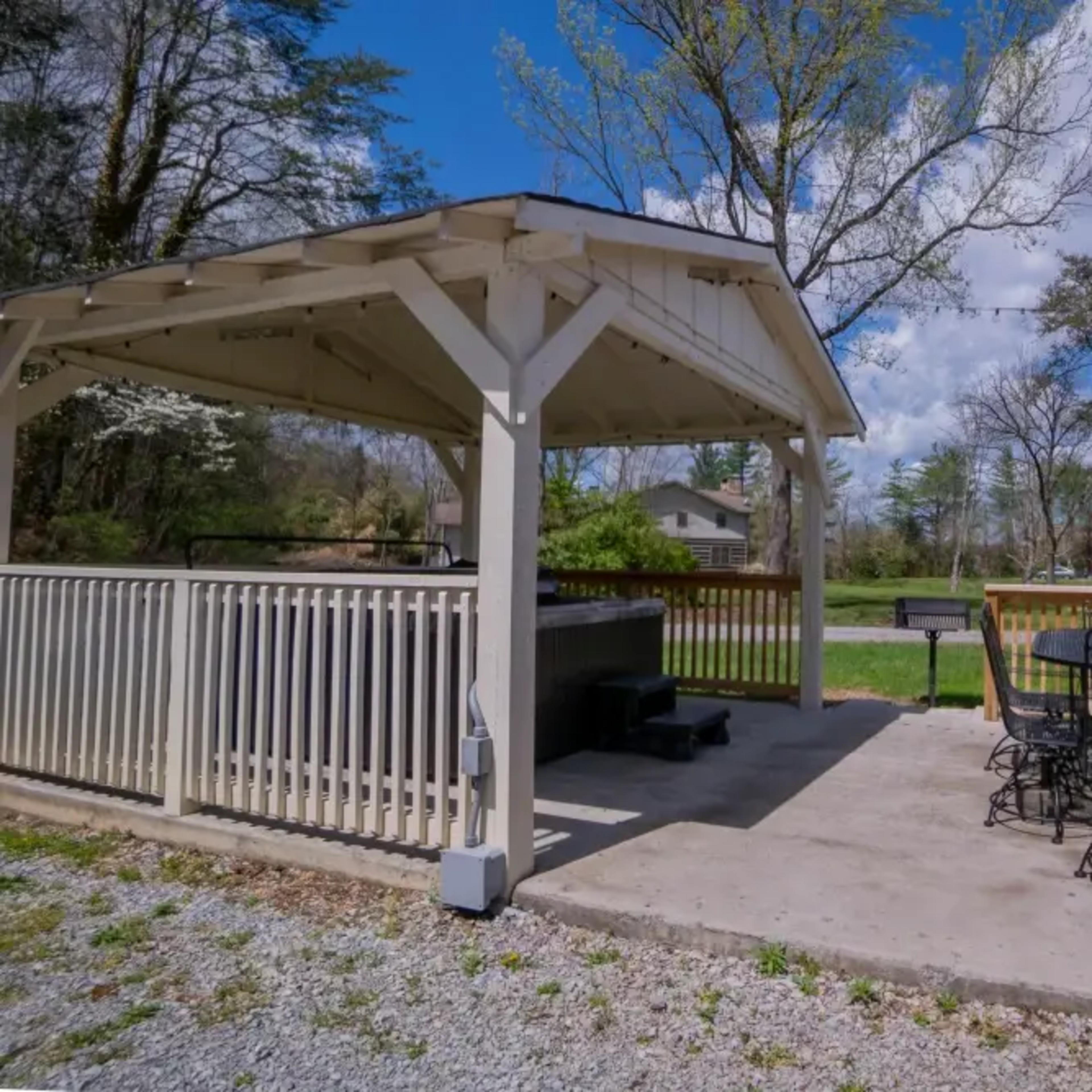 A gazebo with a seating area and a hot tub on a concrete patio, surrounded by trees and grass.