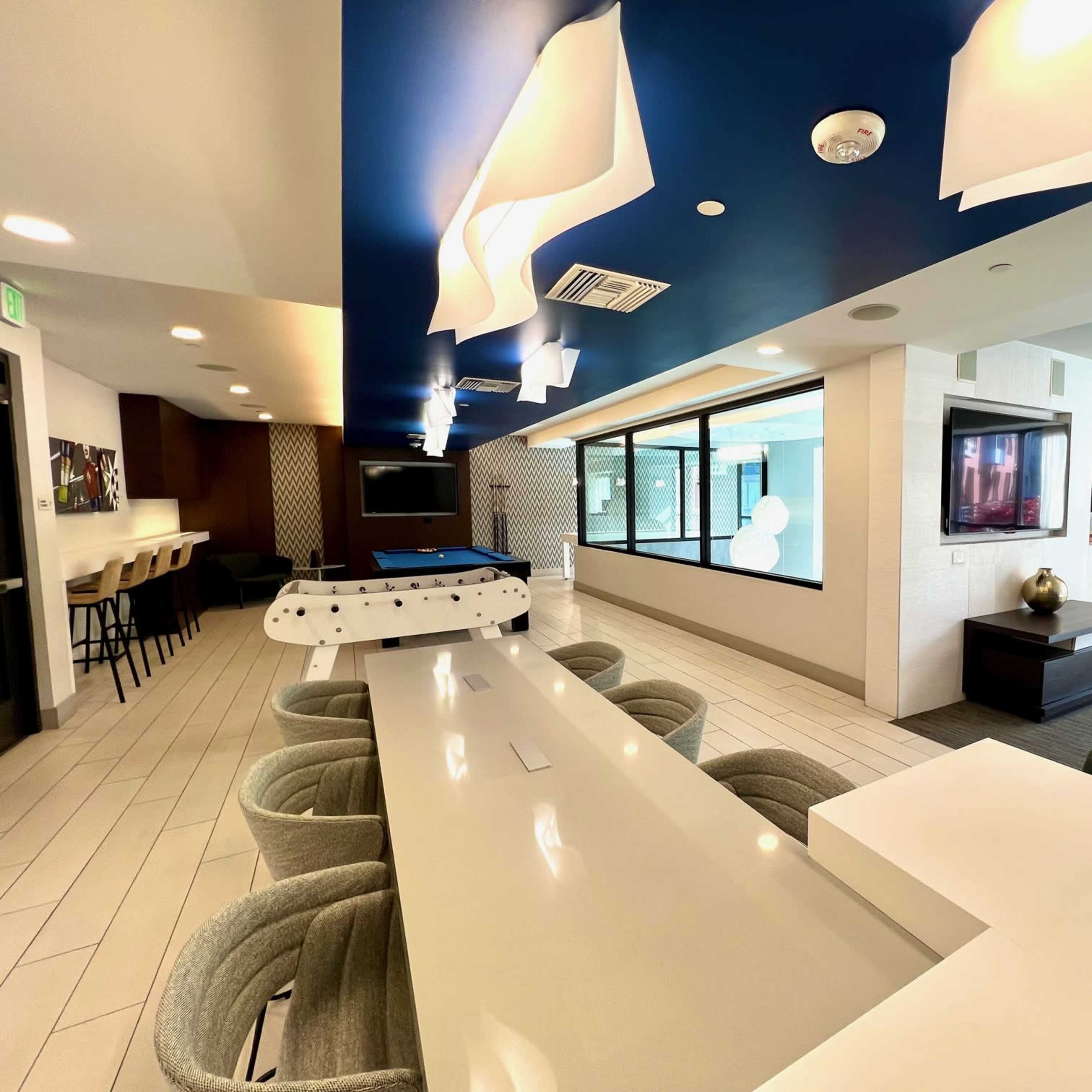 A modern clubhouse area featuring a long white table, bar seating, a pool table, and large windows overlooking a lounge space.