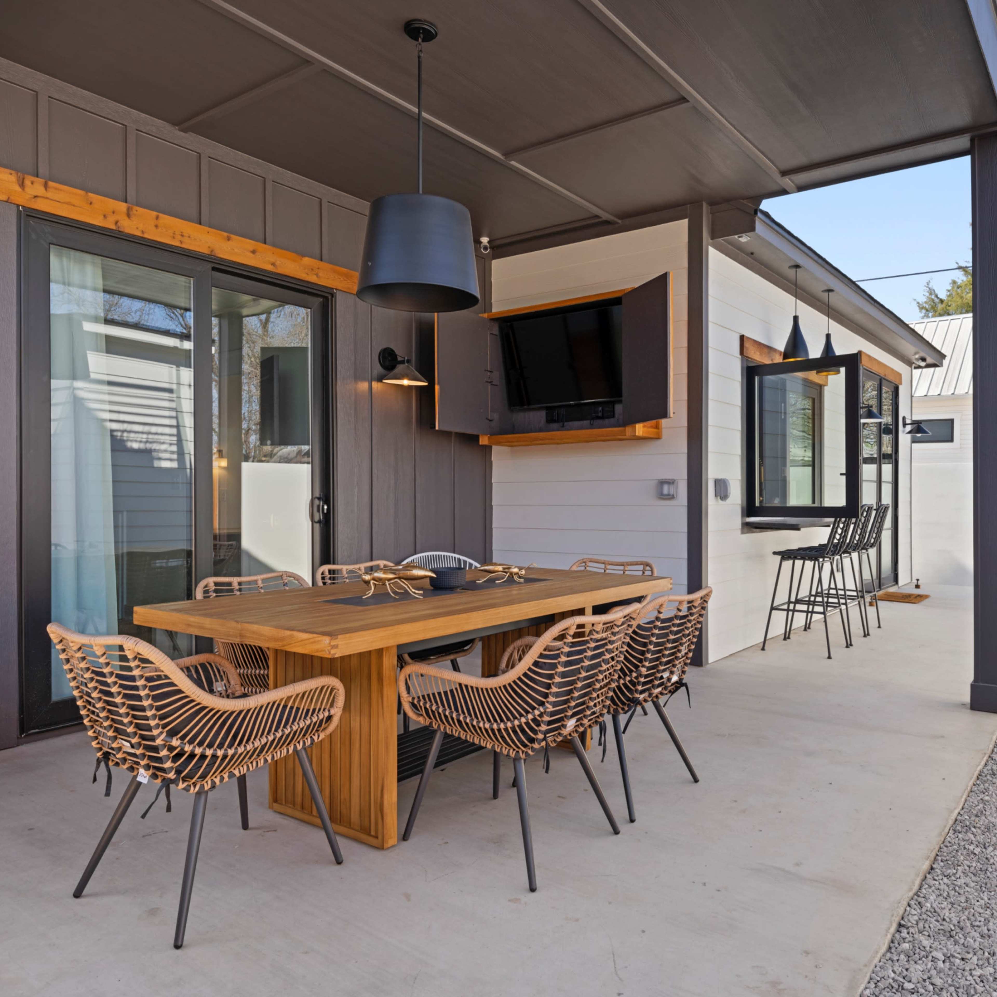 A modern outdoor dining area featuring a wooden table with eight woven chairs, a mounted TV, and a decorative sculpture in a gravel landscape.