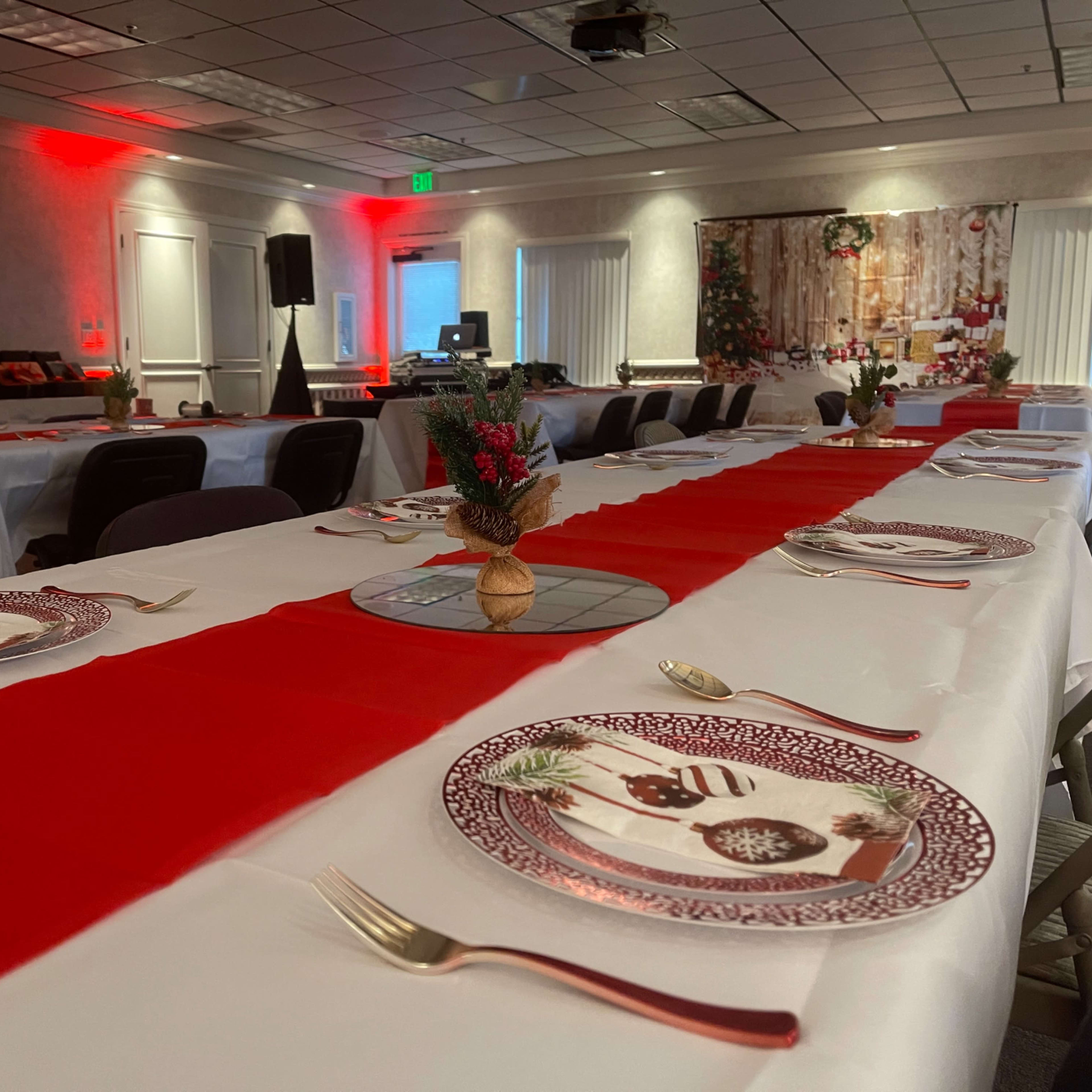A long table set for an event features red table runners, decorative plates, and small floral centerpieces in a well-lit room with soft red lighting.