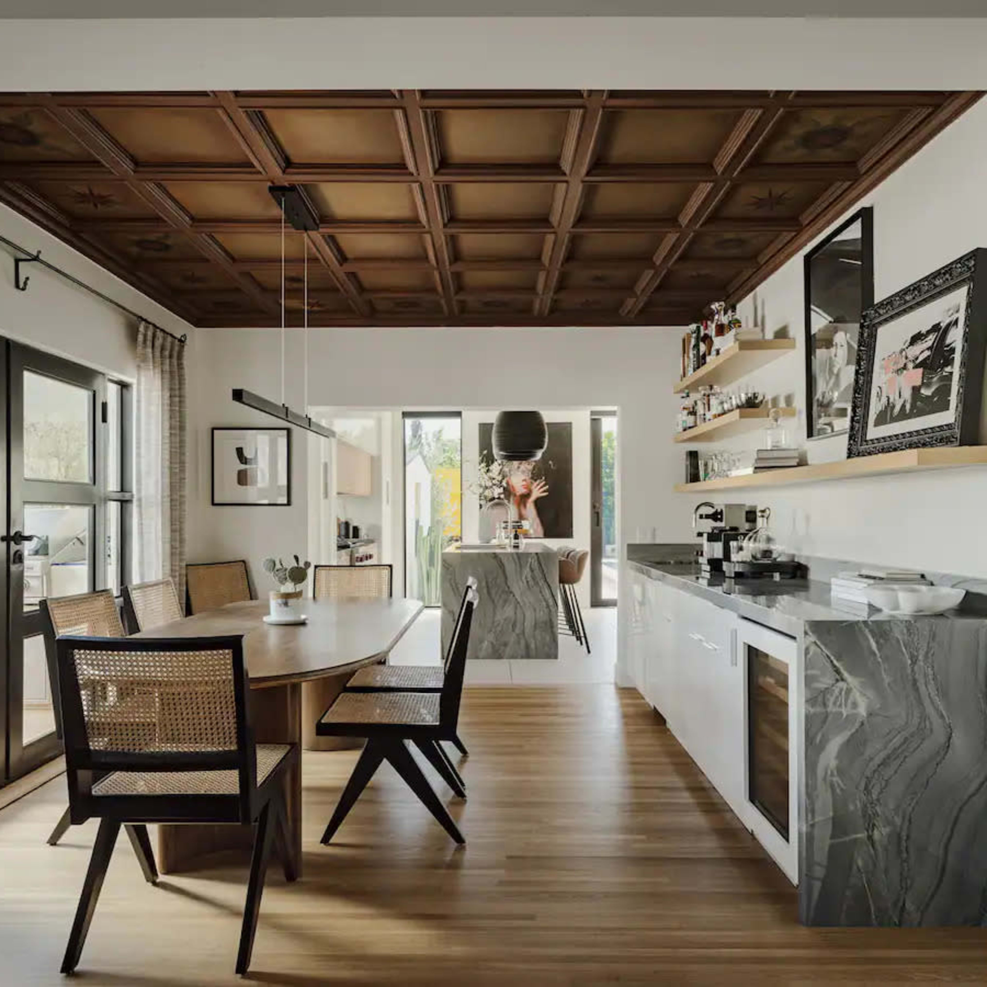 A modern dining area featuring a wooden coffered ceiling, a round table with six chairs, and a sleek kitchen with marble accents and open shelving.