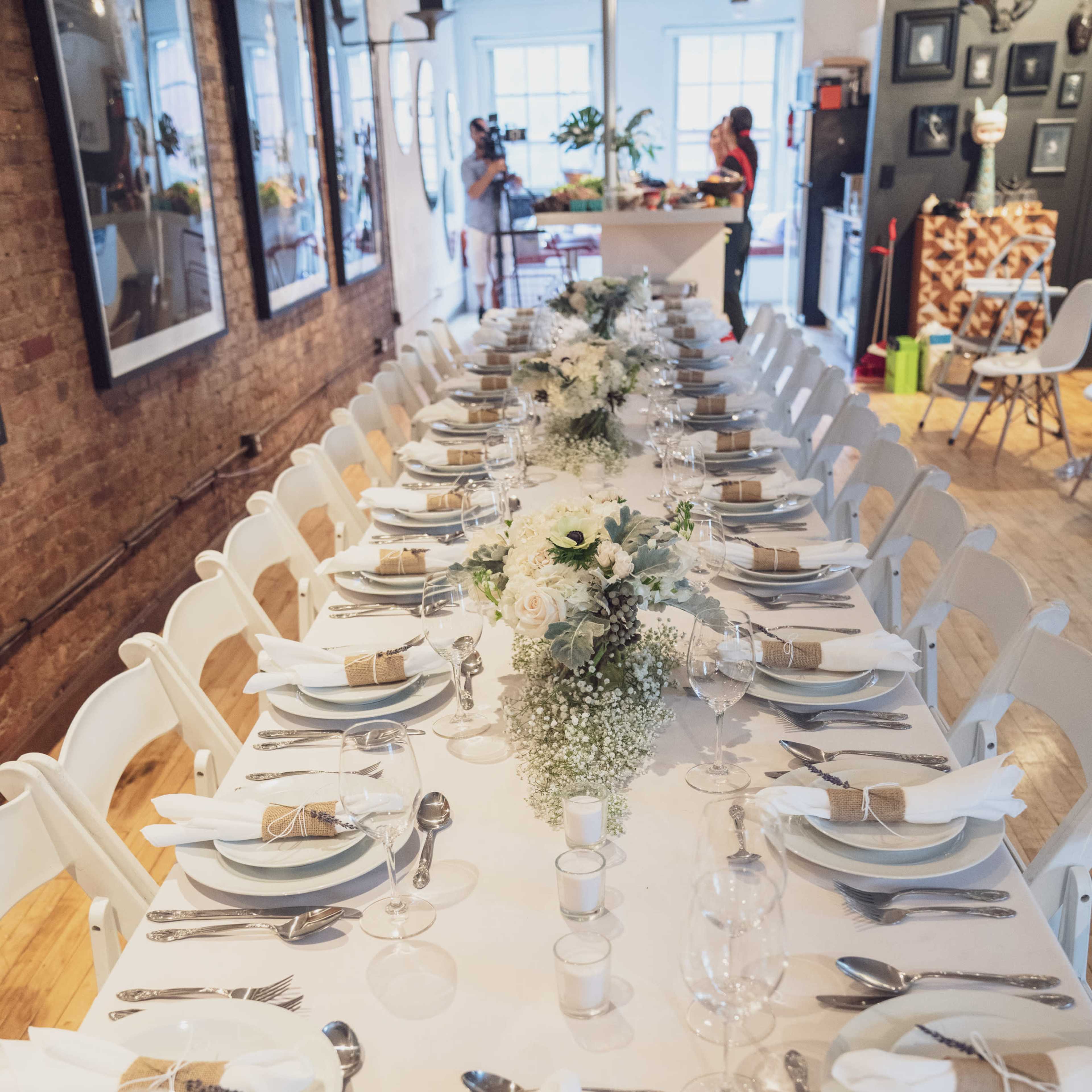 A long dining table set with white tableware and floral centerpieces is arranged in a bright room with brick walls.