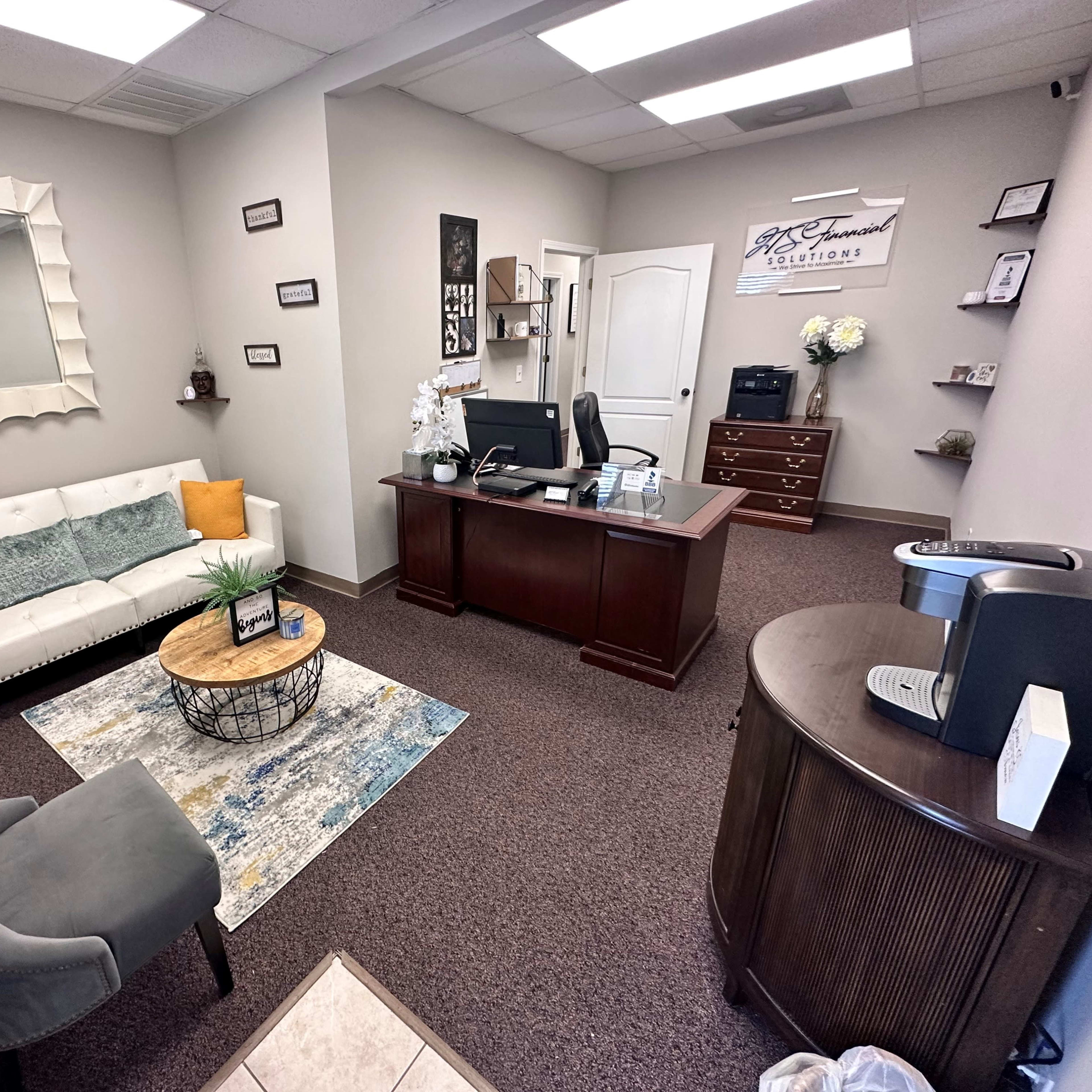 A neatly arranged office with a reception area featuring a white couch, a round coffee table, a desk, and various decorative elements.