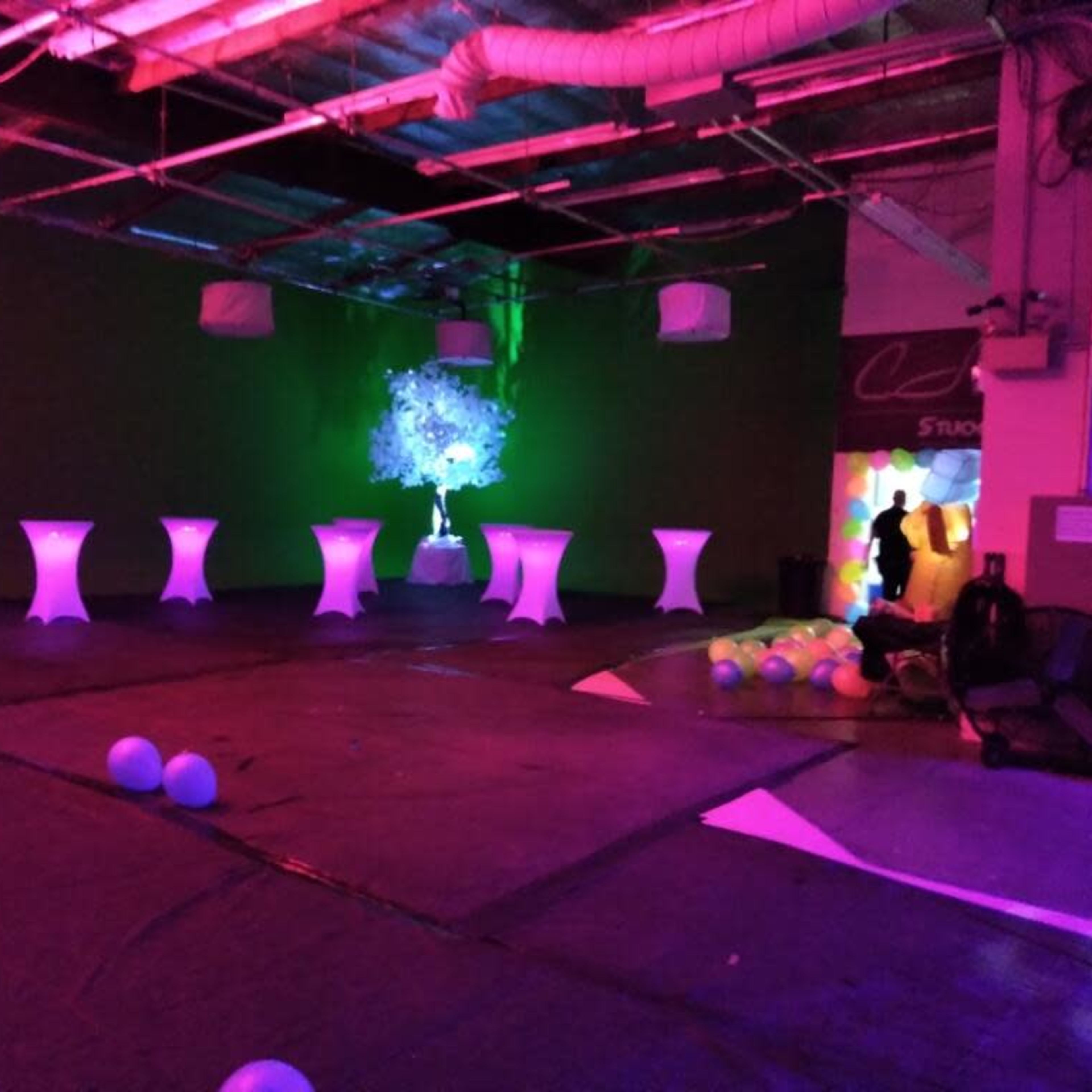 The scene features a dimly lit event space with illuminated tables, a decorative tree, and scattered balloons on the floor.