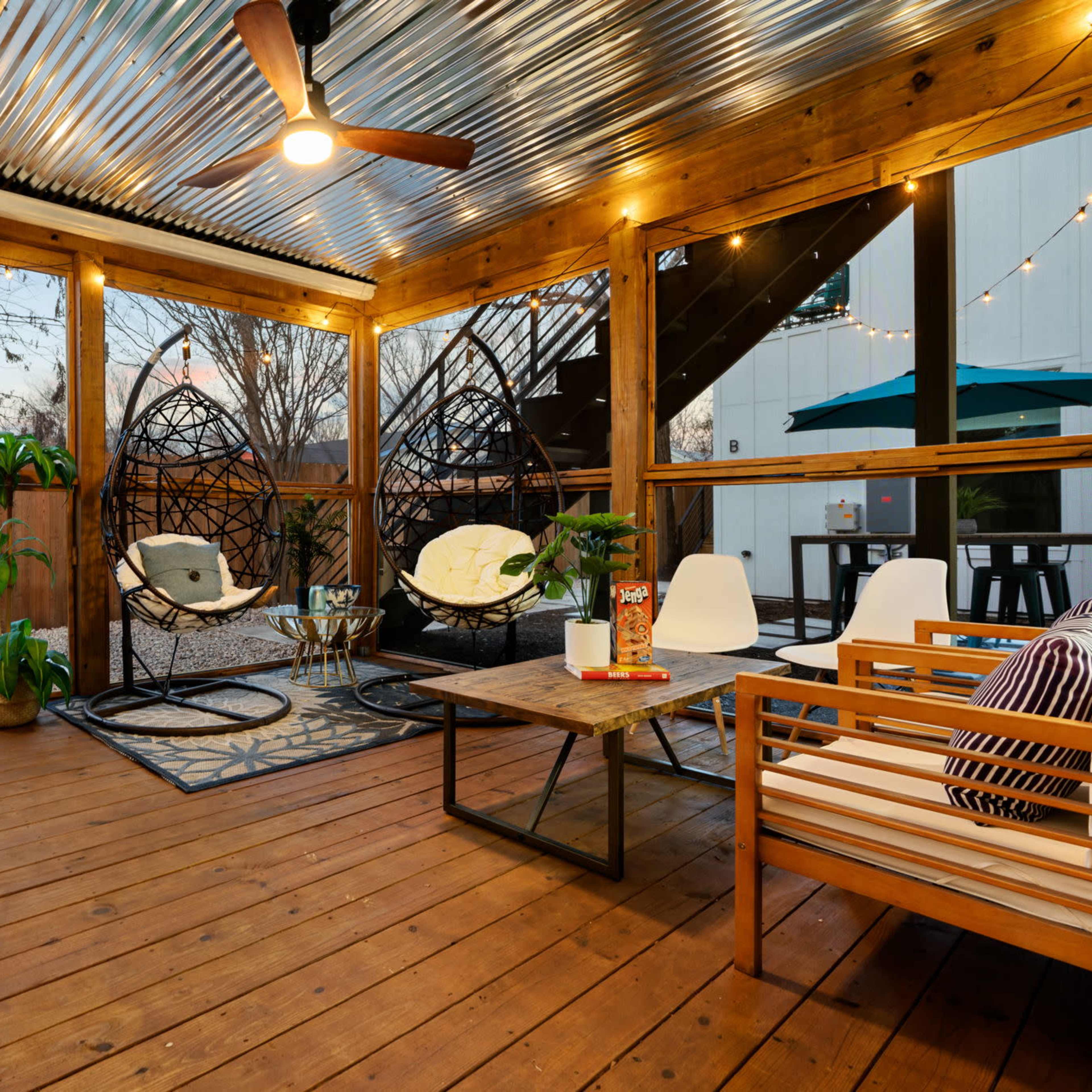 A wooden deck patio featuring a seating area with modern furniture, two hanging chairs, and plants, enclosed by a wooden railing and a corrugated metal roof.